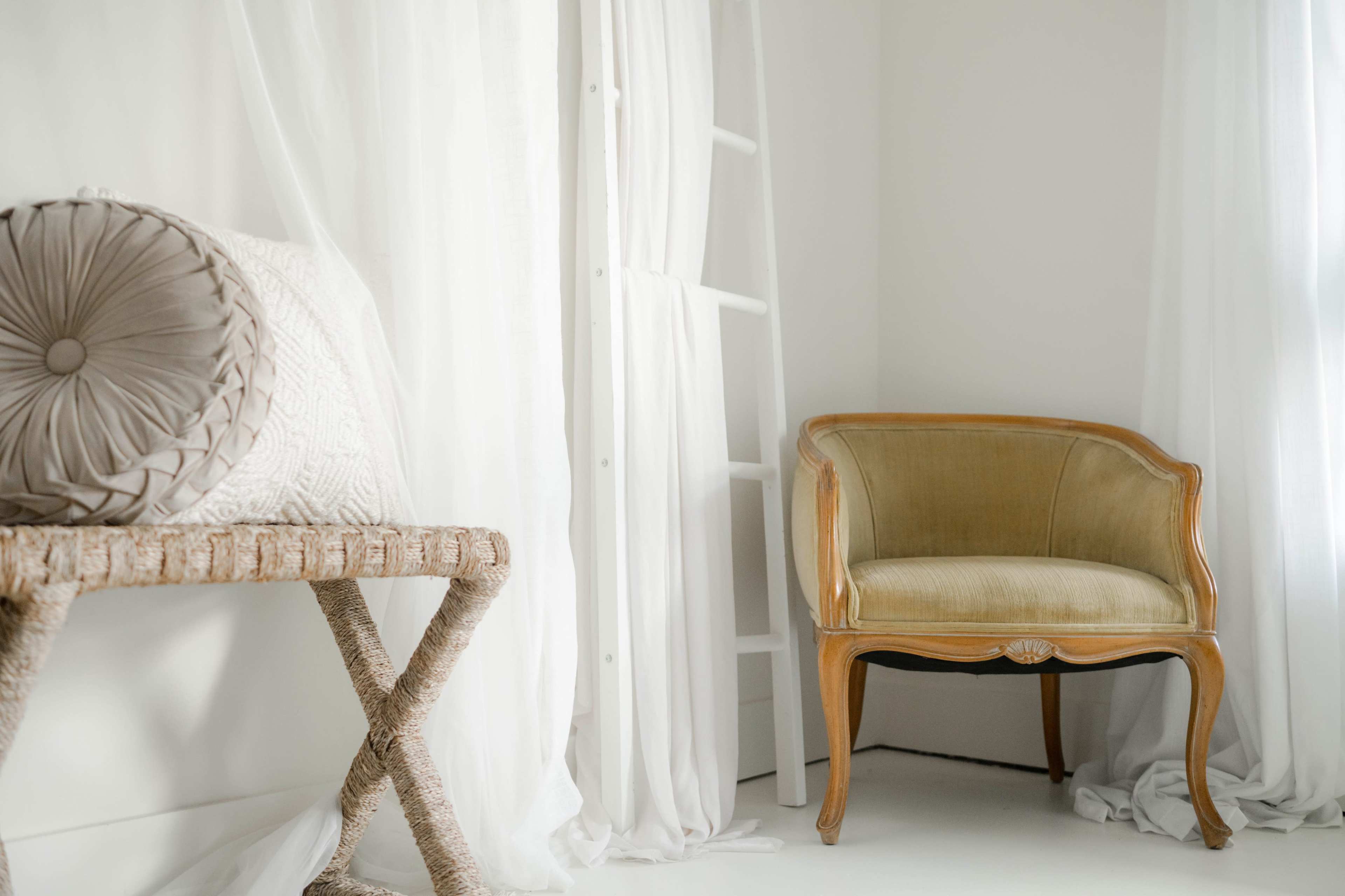 The image shows a minimalist room featuring a wooden chair, a woven side table, and sheer white curtains.