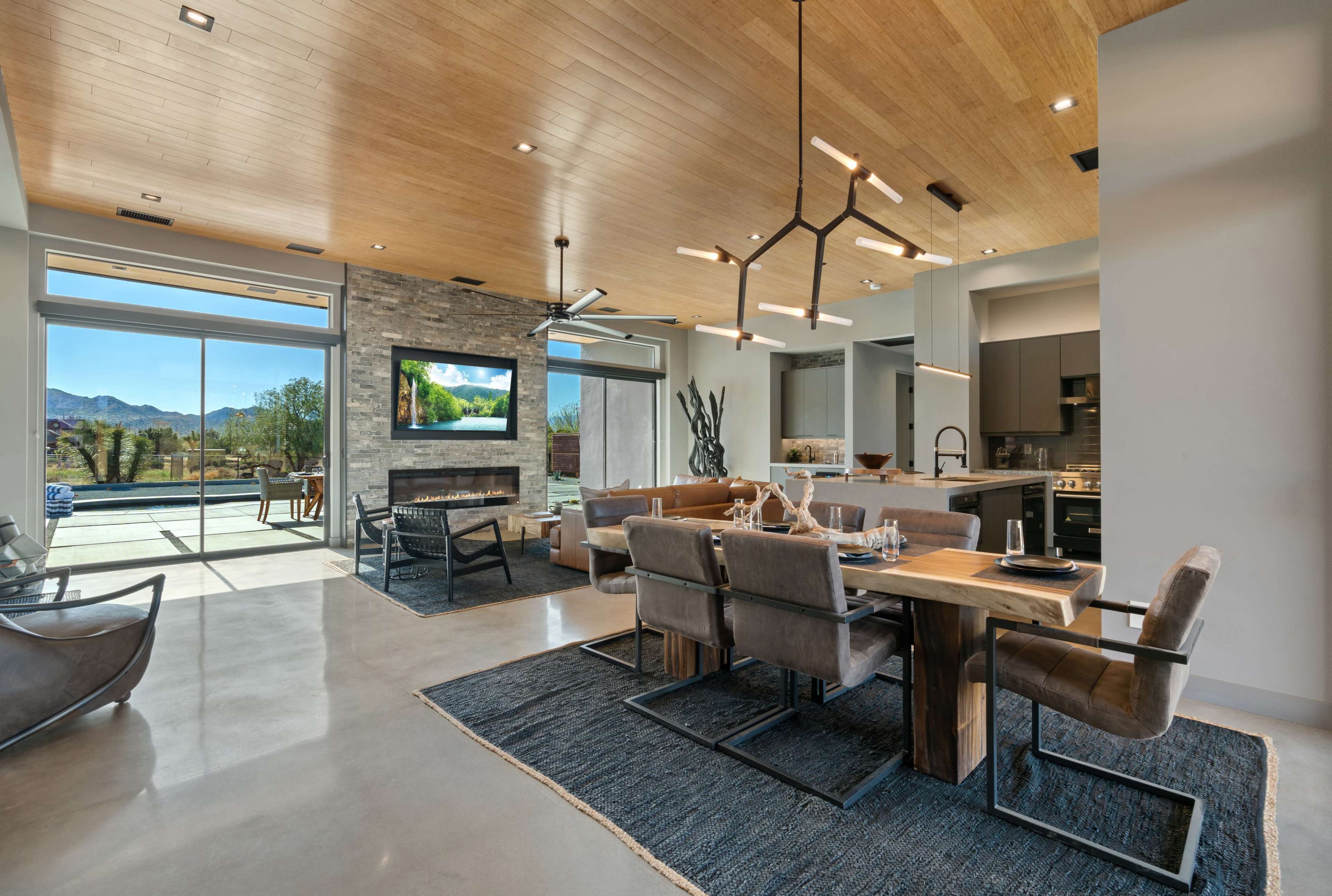 A modern living and dining area features a wooden ceiling, large windows with outdoor views, and a stone fireplace.