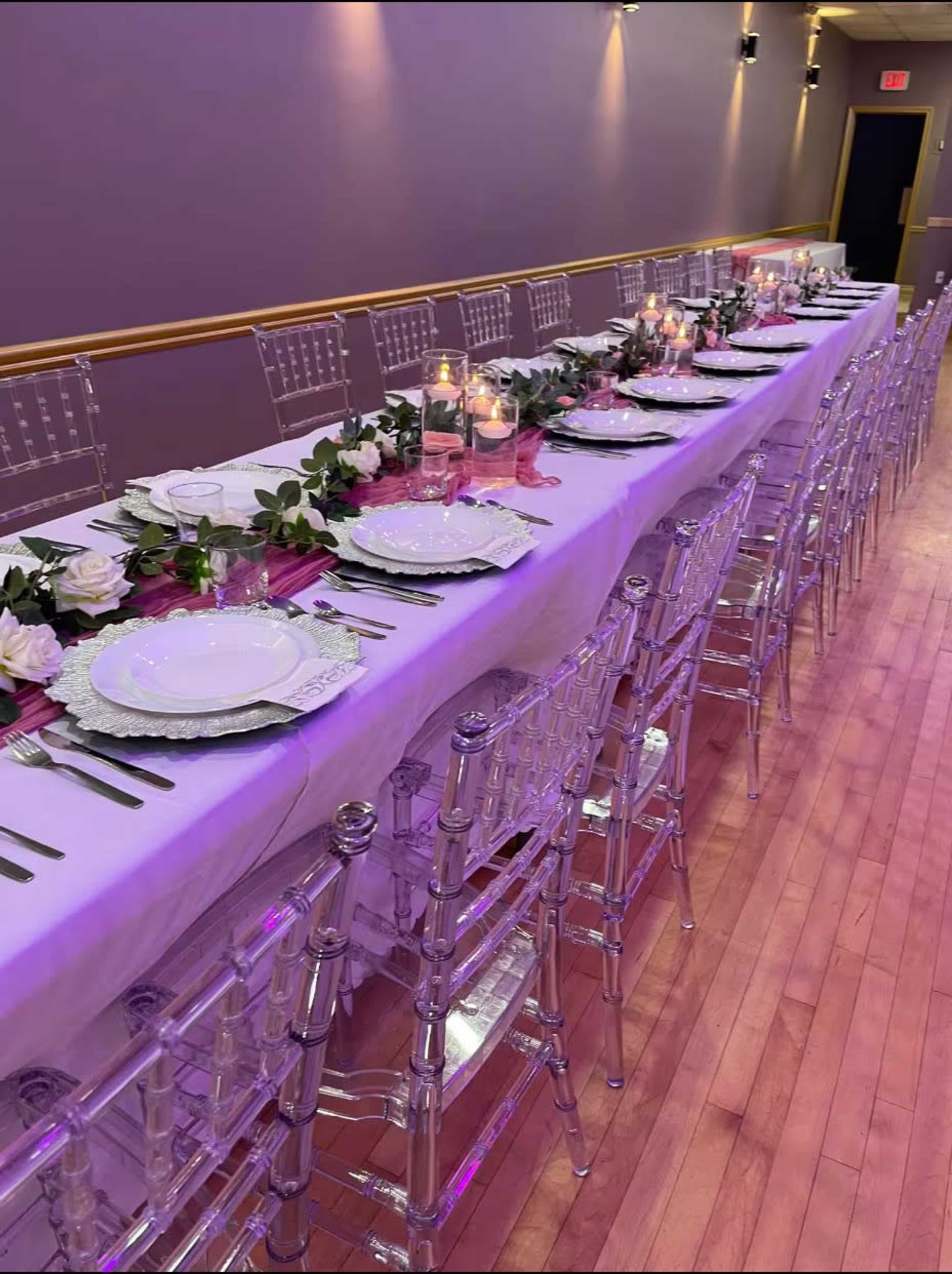 A long banquet table is set with white plates, silverware, and candles, decorated with greenery and white roses, under soft lighting.