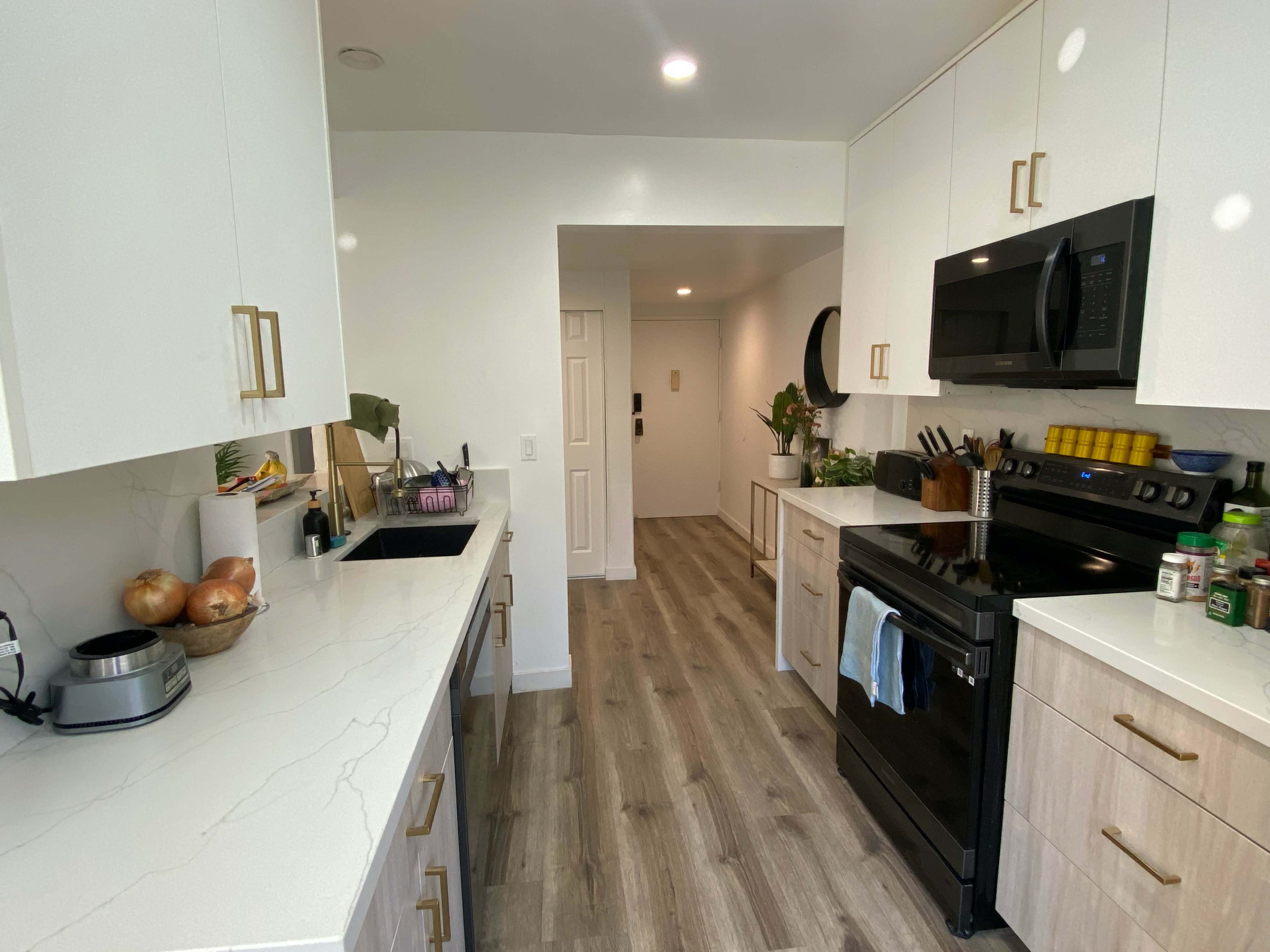 The kitchen features white cabinets, a black stove, and a long countertop with modern appliances and a hallway leading to a door at the end.