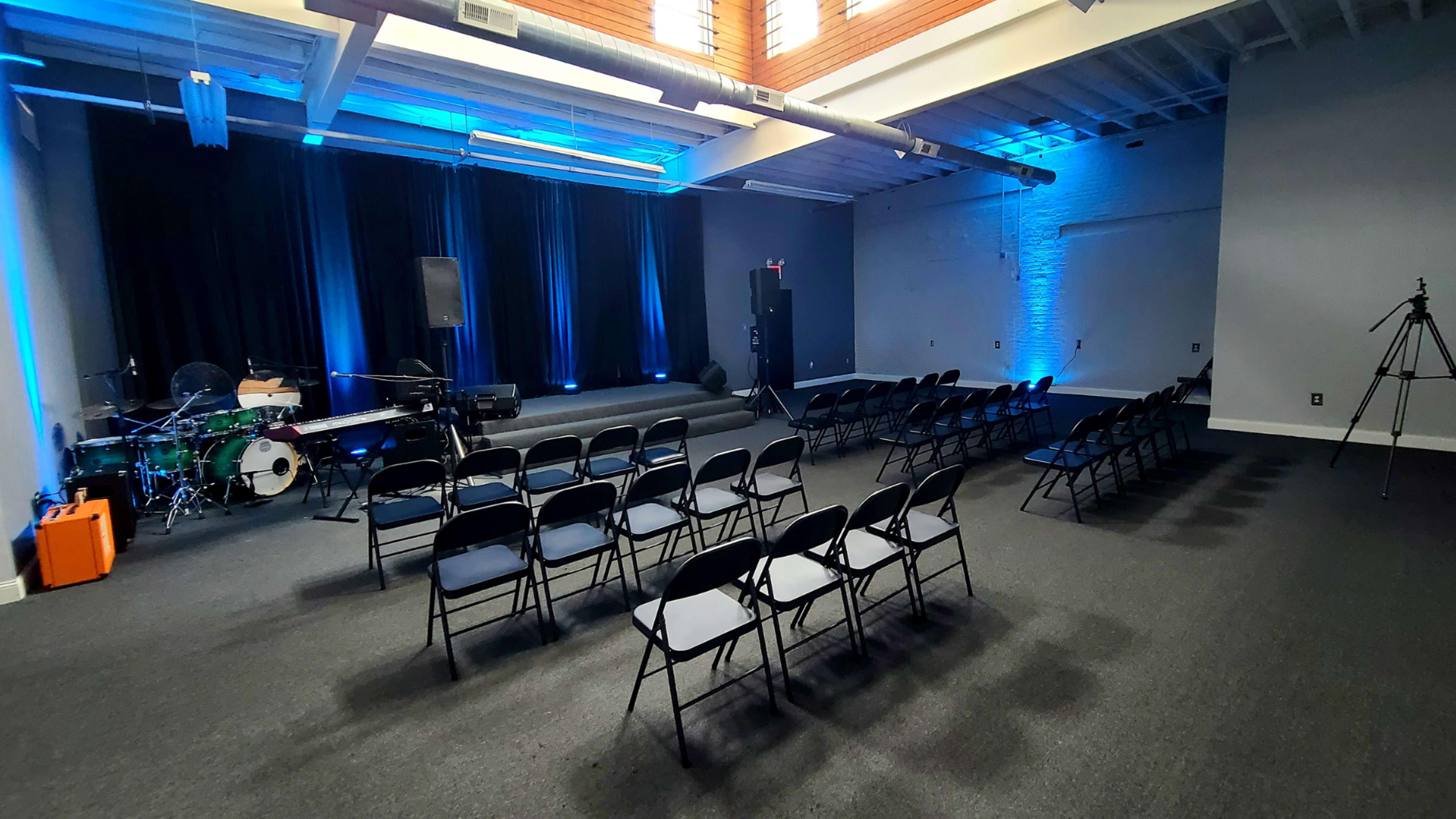 A performance space with black chairs arranged in rows facing a small stage, flanked by blue-lit curtains and a drum set in the corner.