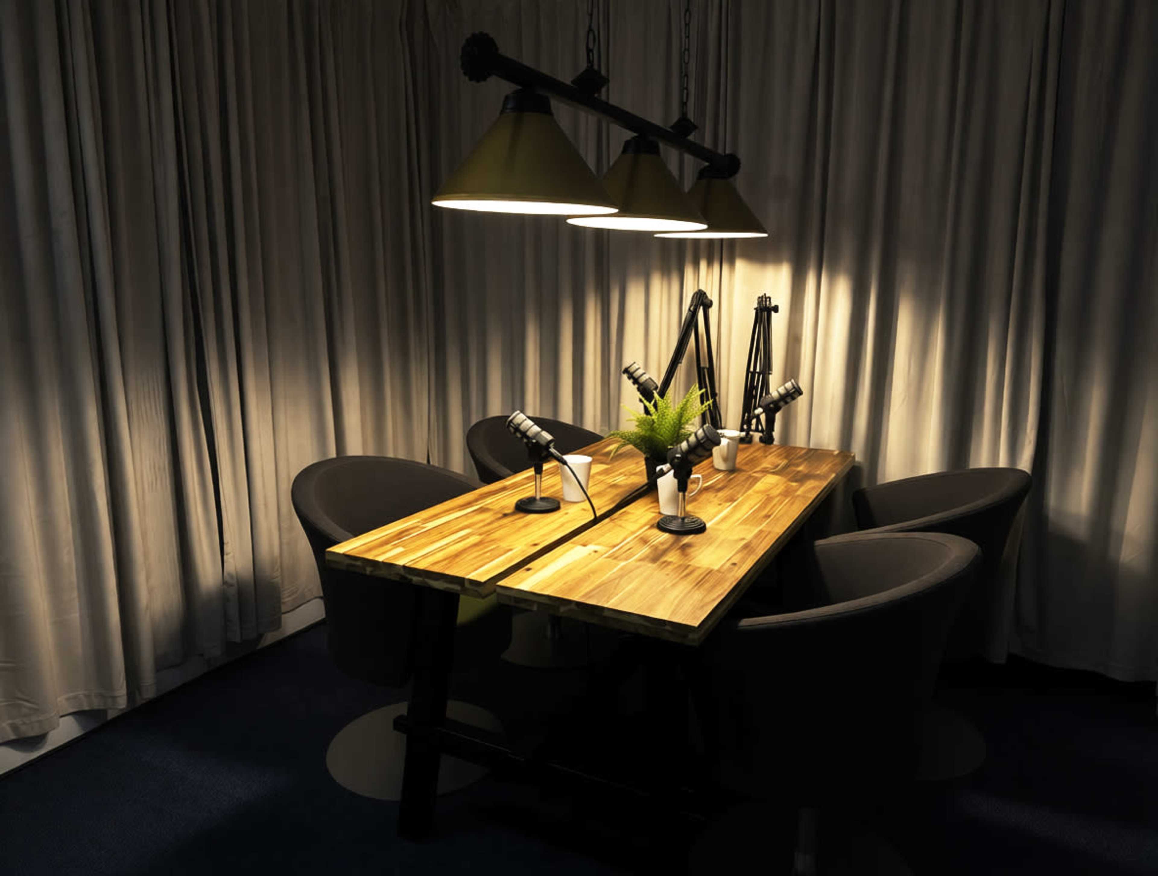 A dimly lit meeting room features a wooden table with microphones and four chairs arranged around it, under pendant lights and a backdrop of curtains.