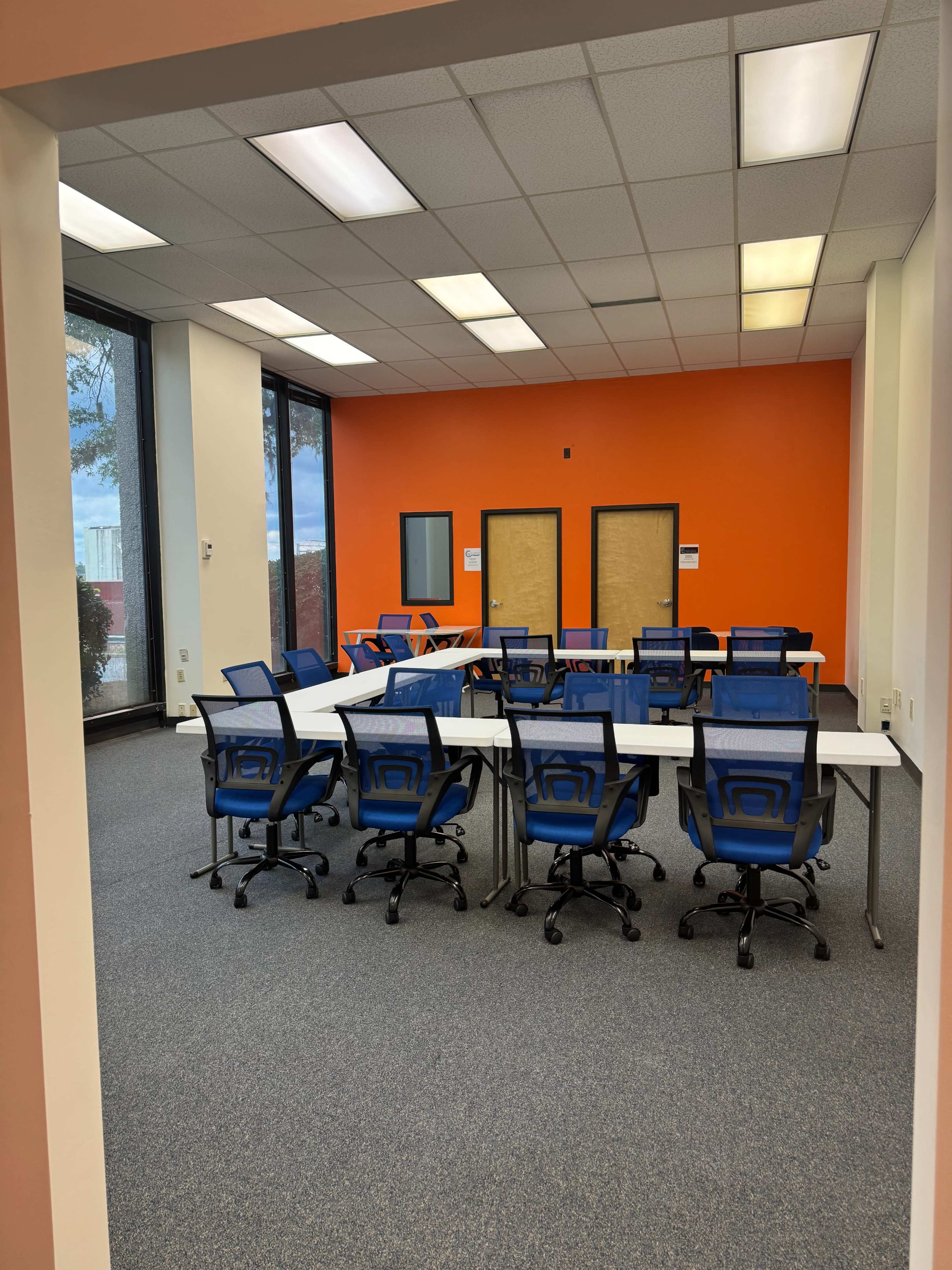 A classroom with several rows of blue chairs and tables arranged in a U-shape, against an orange wall with two closed doors.
