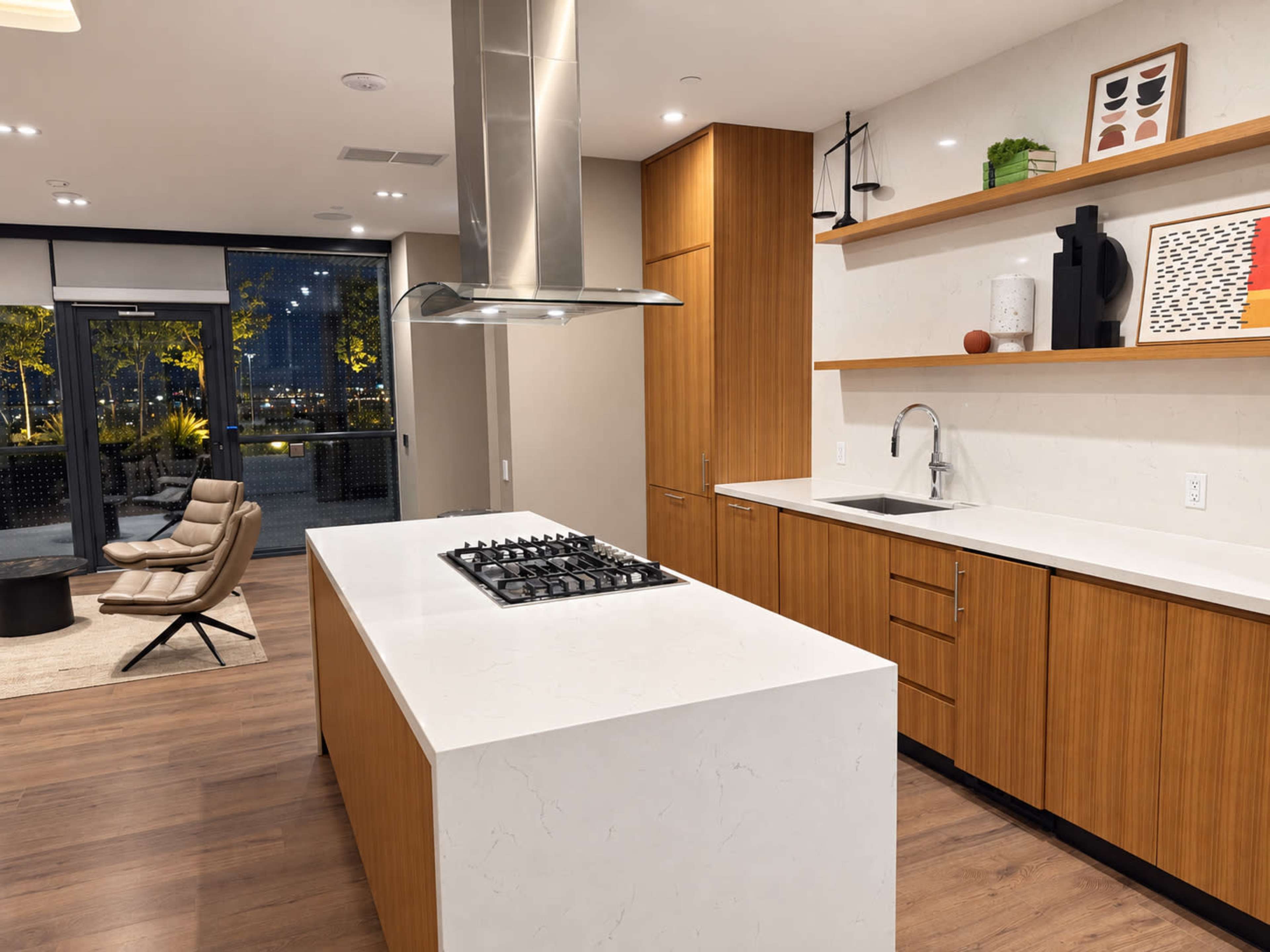A modern kitchen features sleek wooden cabinets, a central island with a gas cooktop, and an adjacent dining area with a large window and outdoor view.