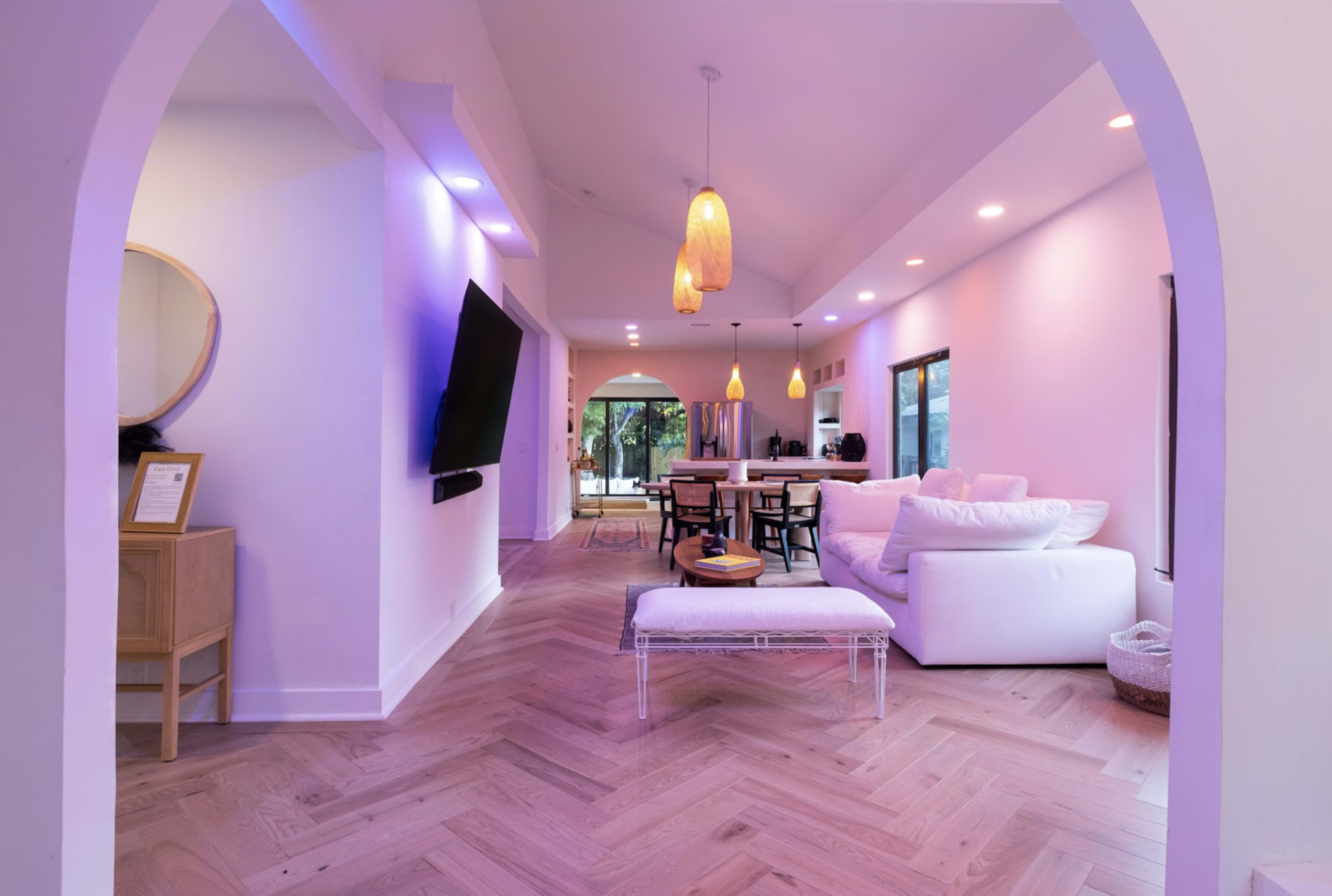 The image shows a modern living space with a white sofa, wooden dining table, and large windows allowing natural light.