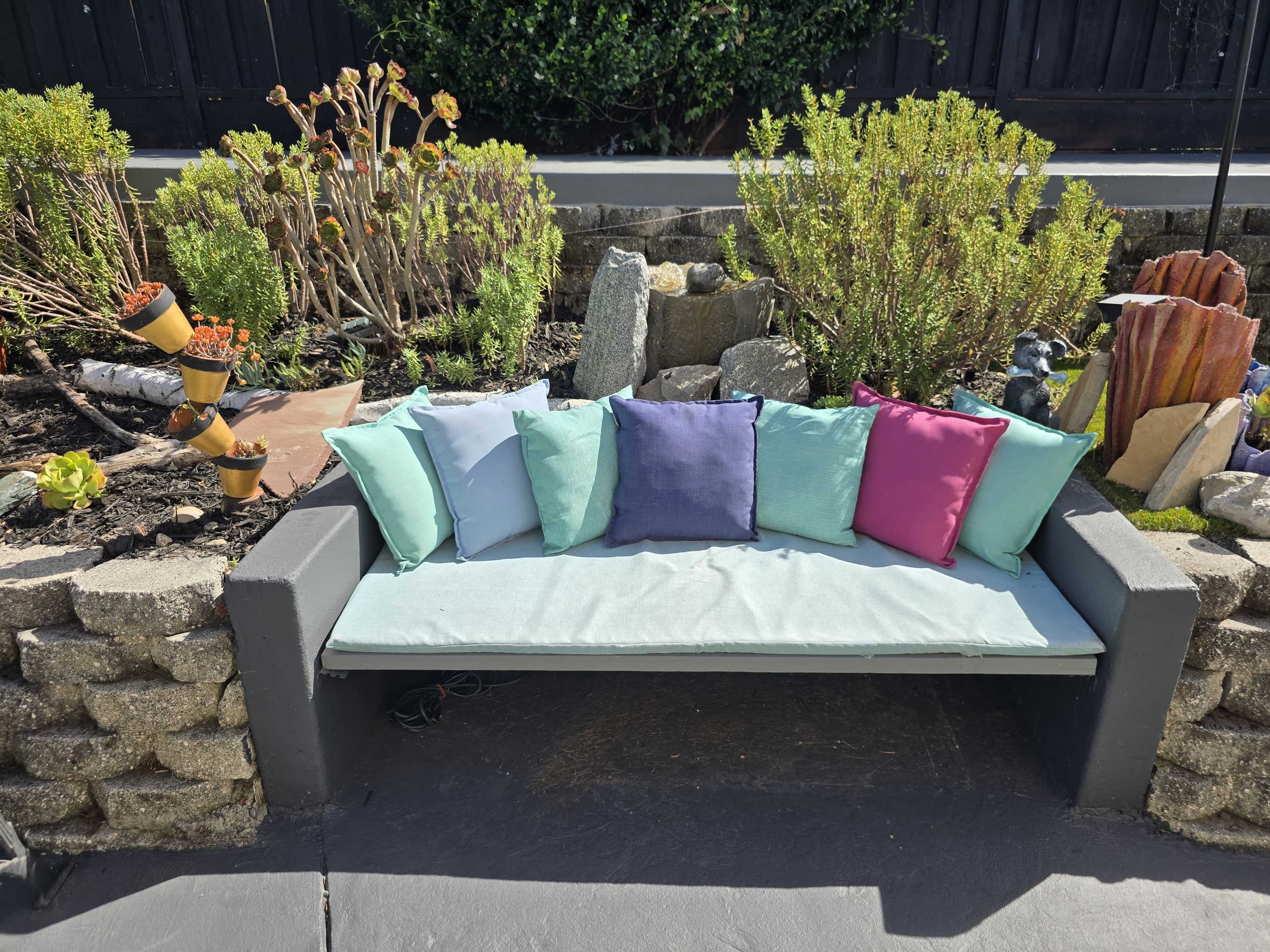 A modern outdoor couch with colorful cushions is positioned among various plants and decorative stones in a landscaped garden.