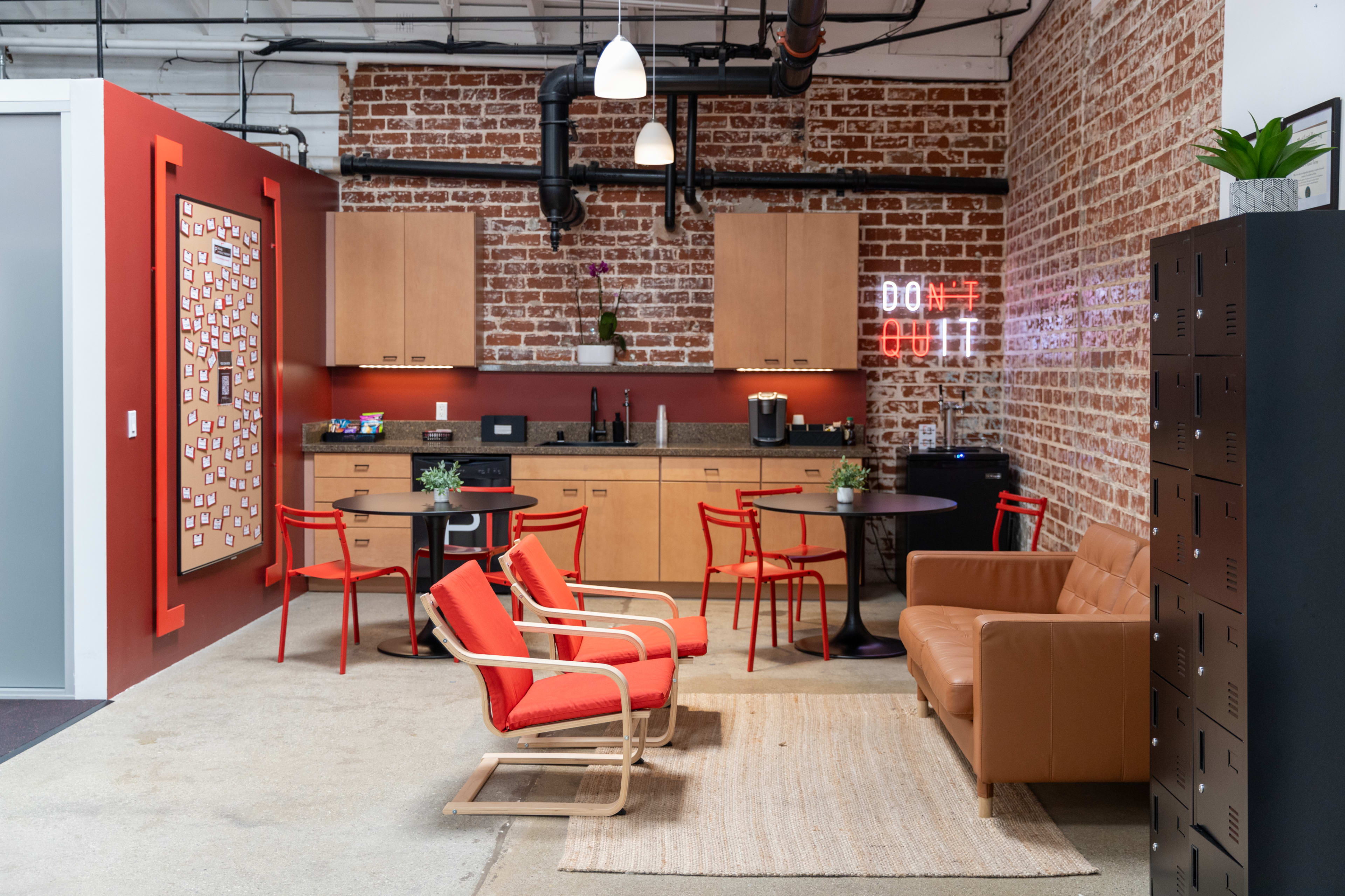 The image shows a modern office break room featuring a kitchenette, seating area with orange chairs and a brown couch, and decorative elements on the walls.