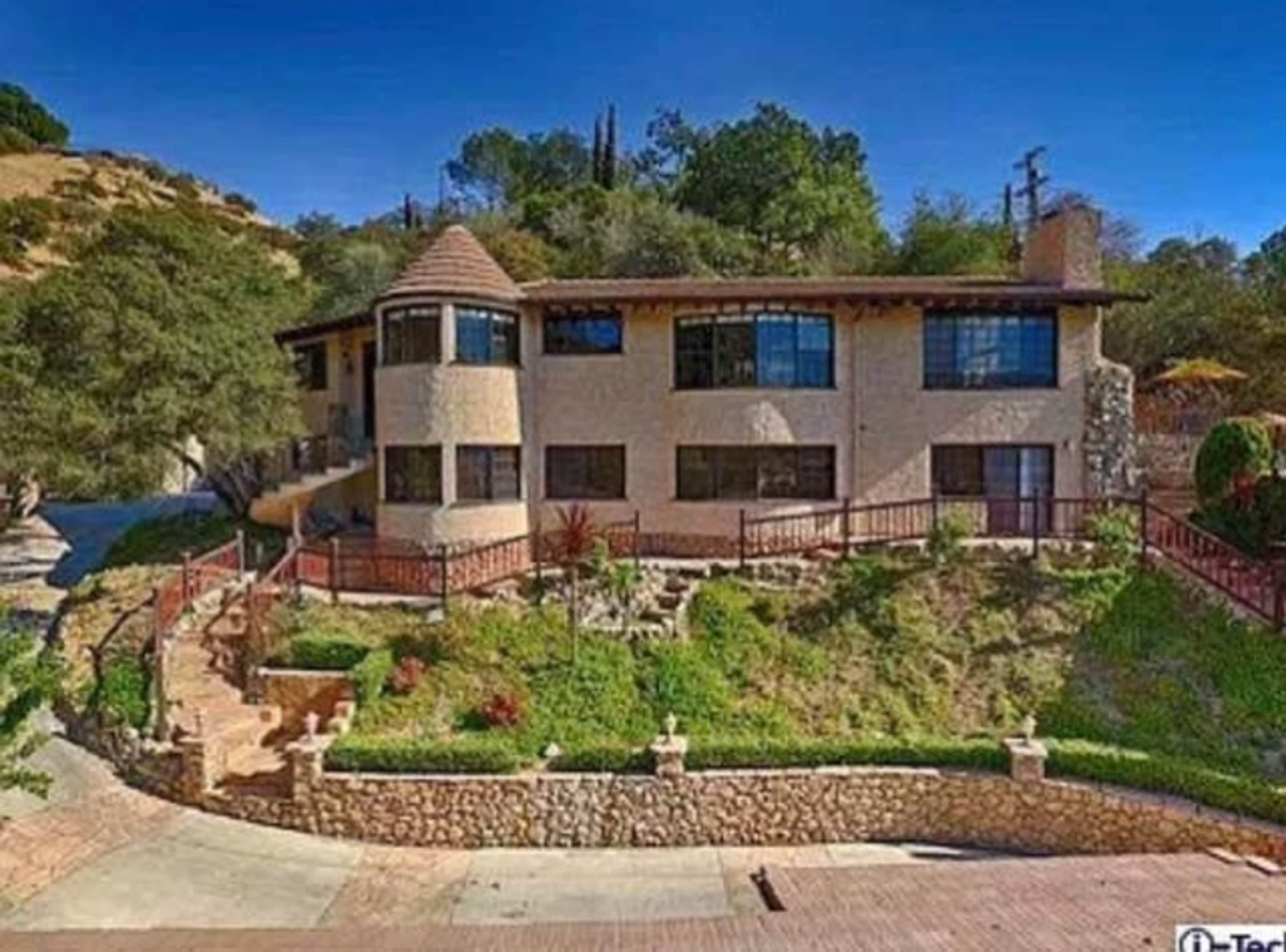 The image shows a multi-level house with a circular turret, surrounded by greenery and a stone pathway.