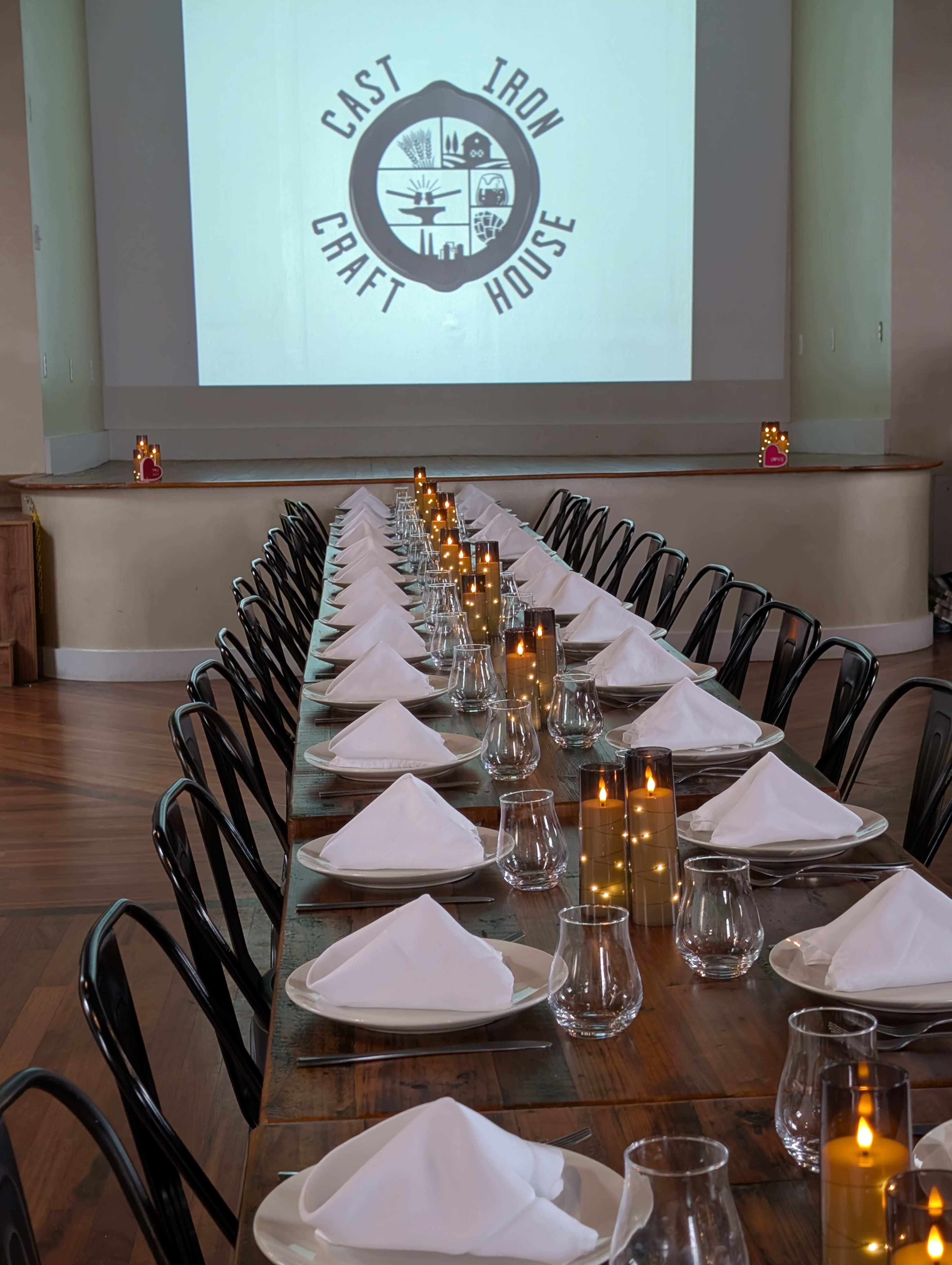 A long banquet table is set with white napkins and glassware, with a logo for "Cast Iron Craft House" displayed on a screen in the background.