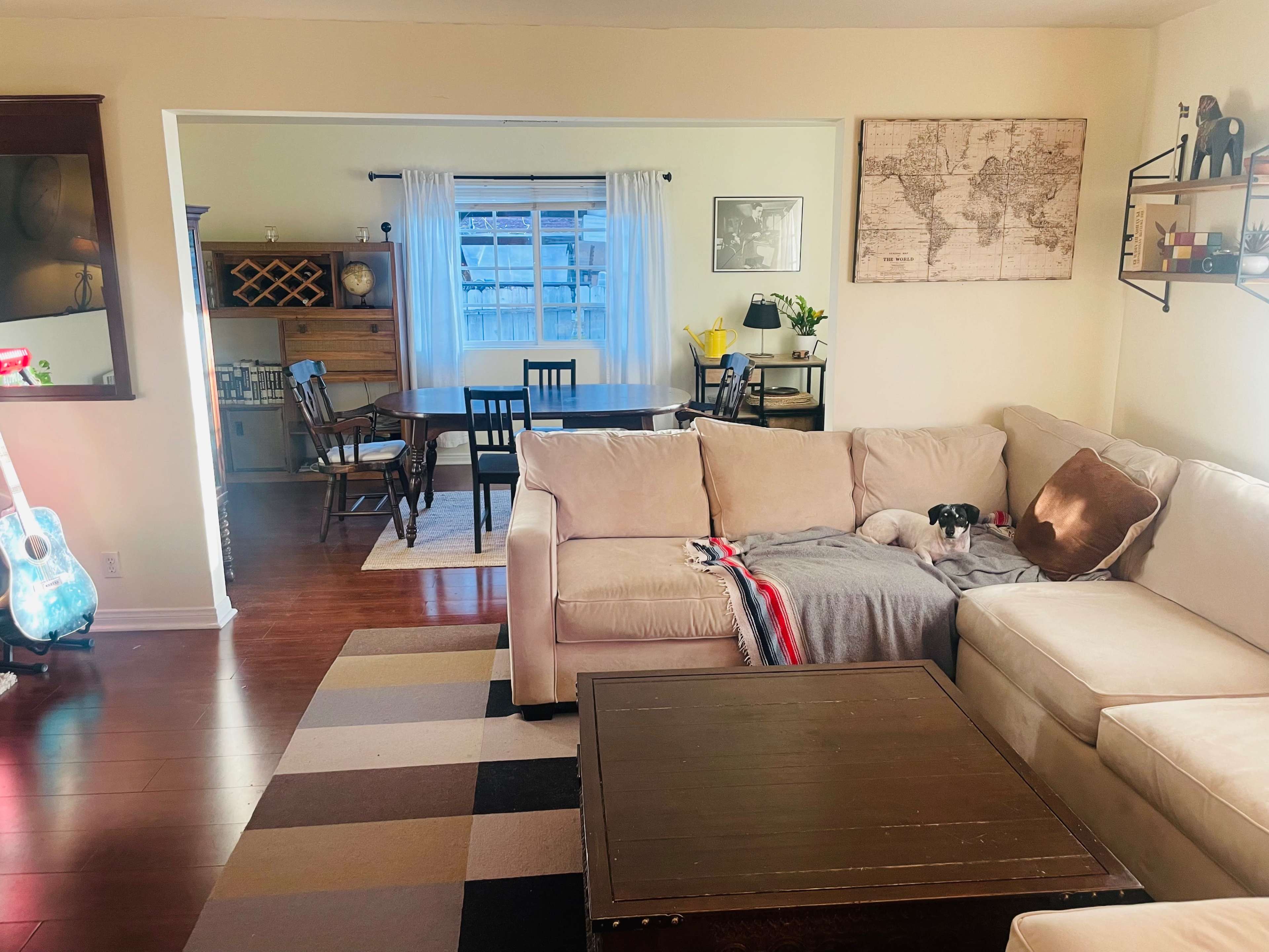 A living room features a beige sectional sofa with a dog resting on a blanket, a coffee table, and a view into a dining area with a wooden table and a wine rack.