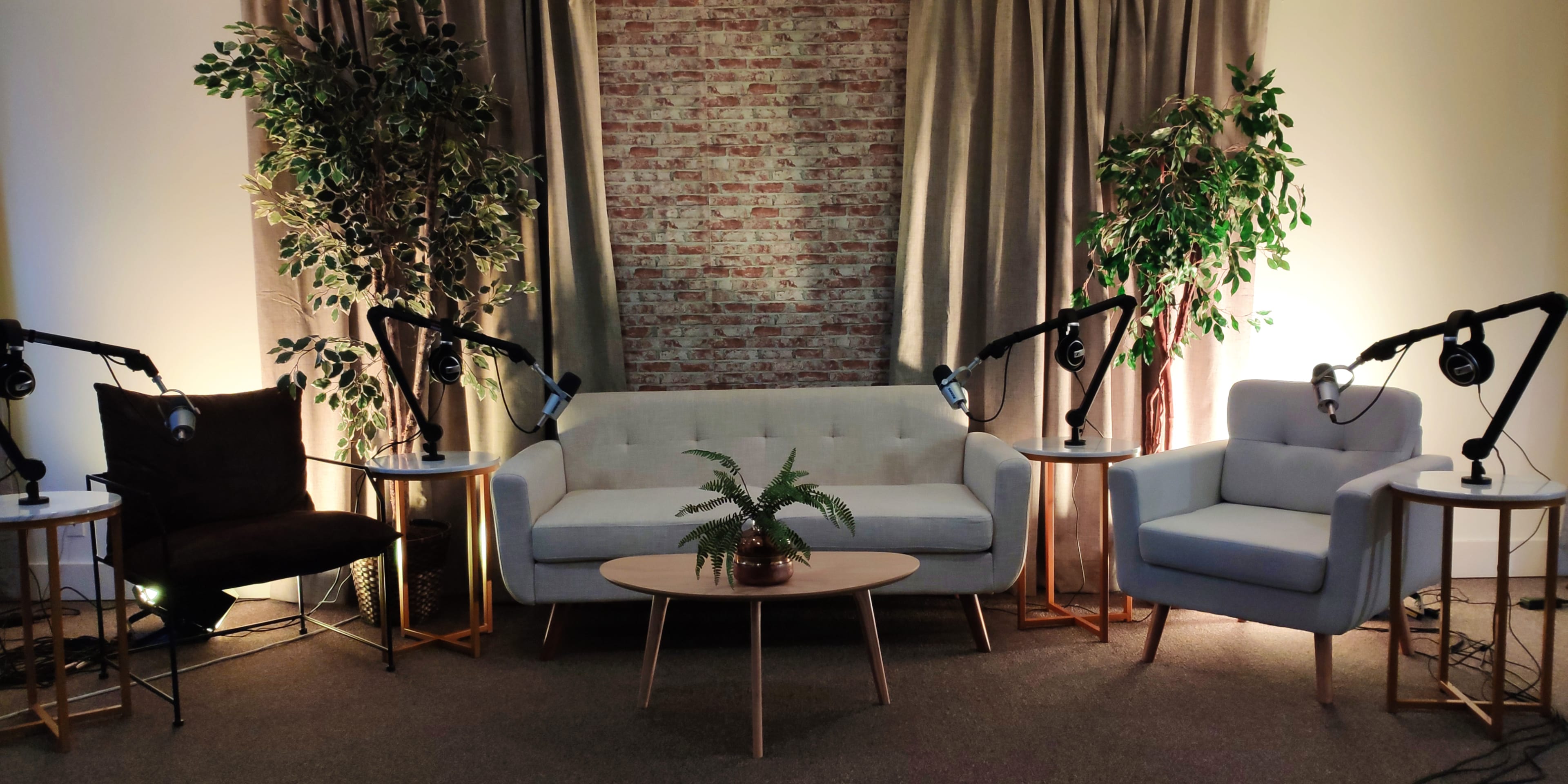 A cozy interview setting with a light gray sofa, two accent chairs, a round coffee table, and several bright floor lamps, surrounded by potted plants against a brick wall backdrop.