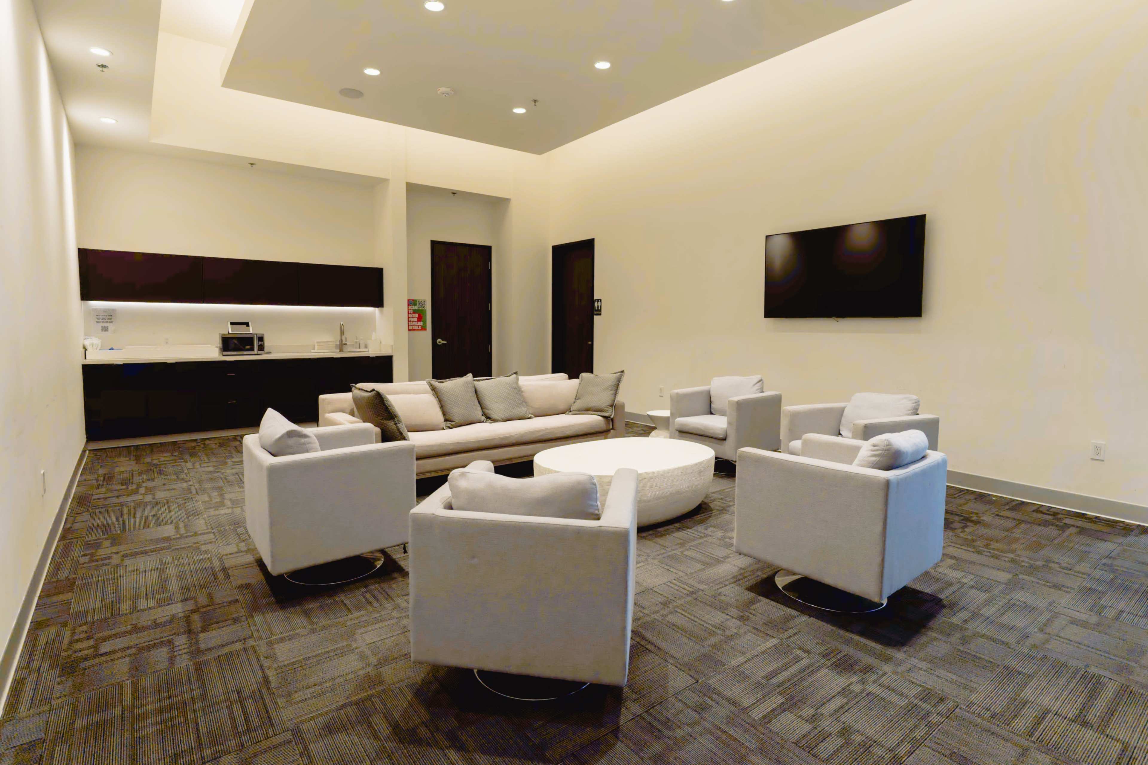 The image shows a modern lounge area with a sofa, circular coffee table, and four armchairs arranged around it, complemented by a wall-mounted TV and a kitchenette in the background.