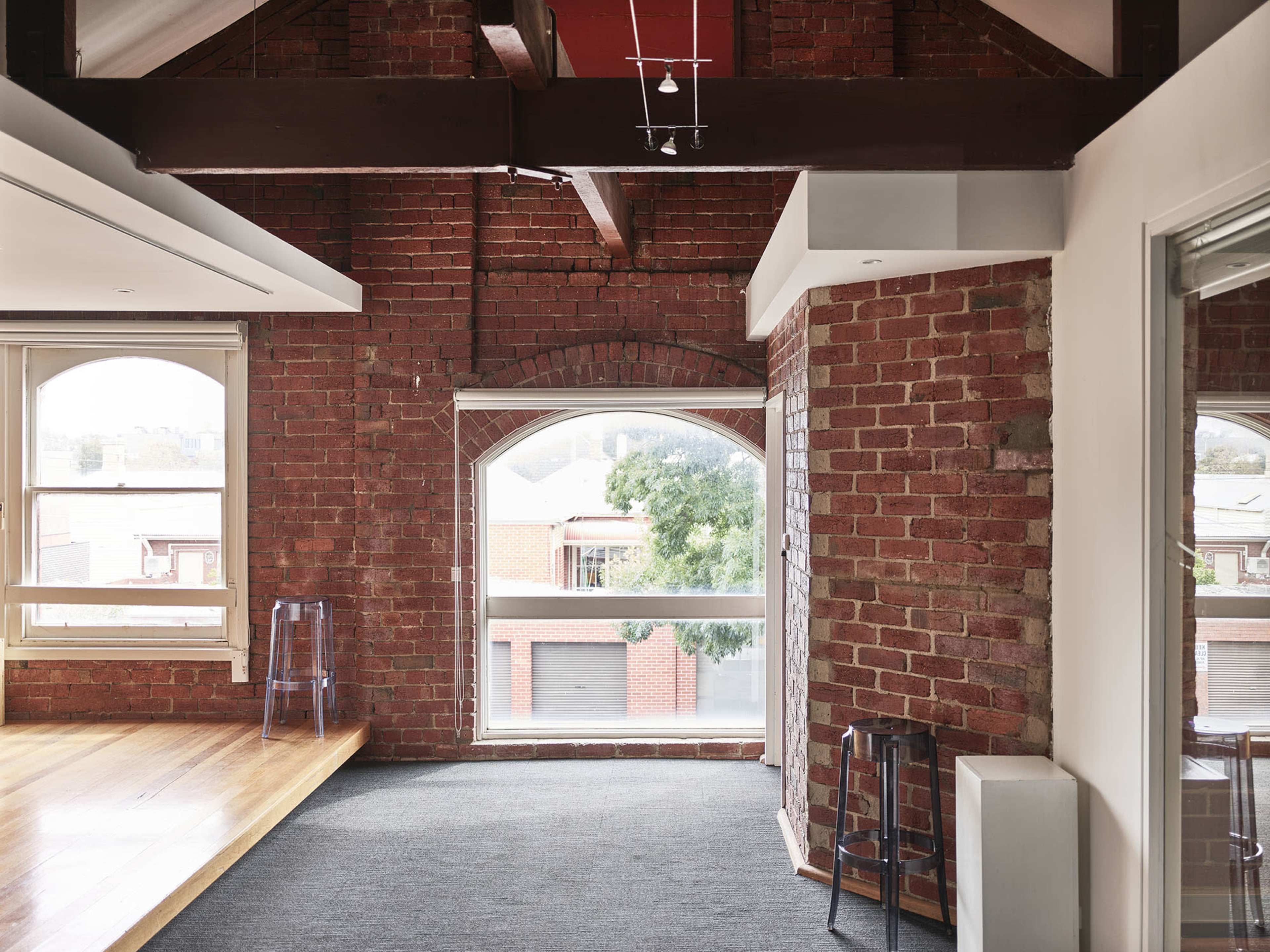An interior space with exposed red brick walls, large arched windows, wooden flooring, and minimal furniture.