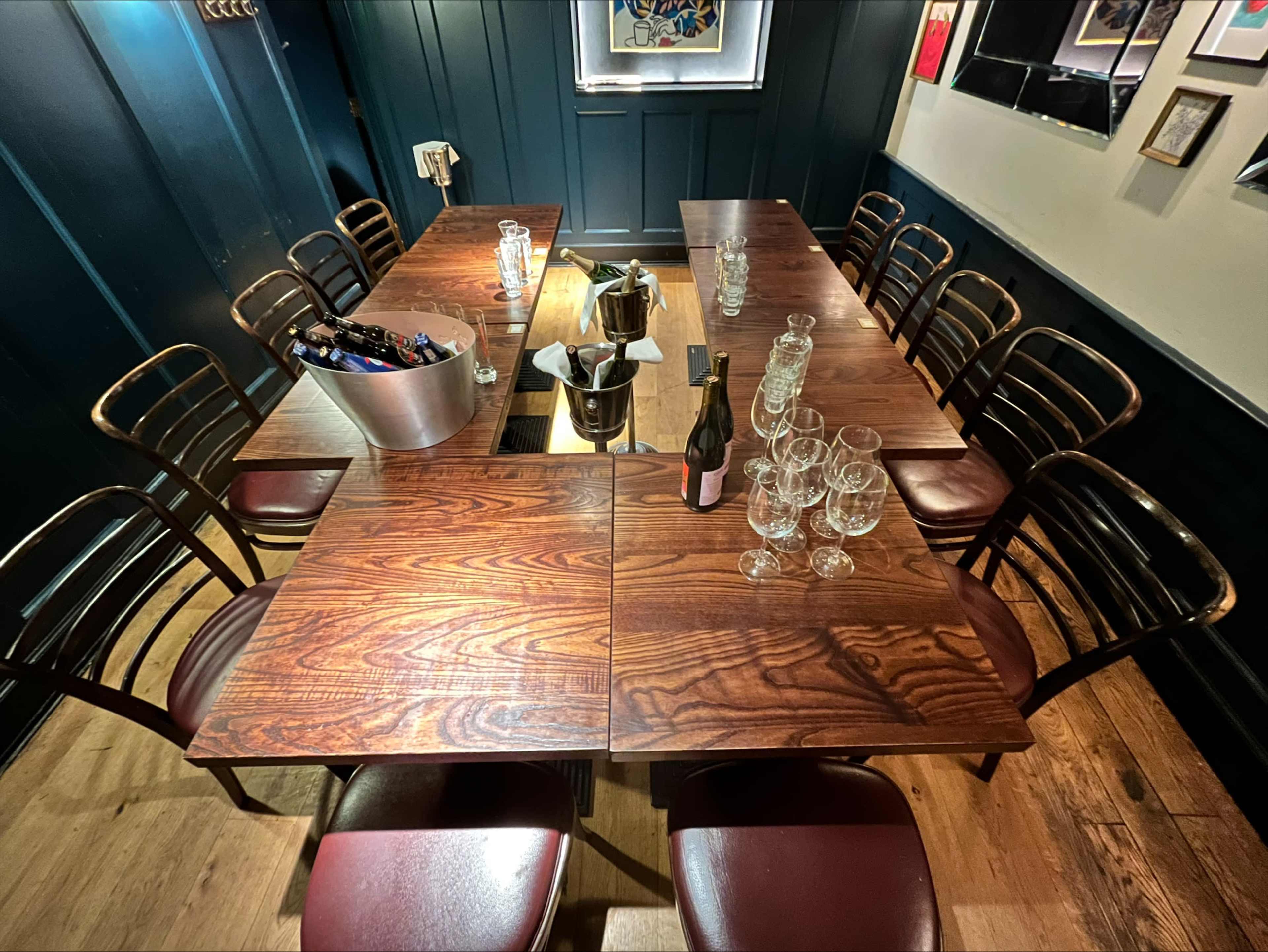 A large wooden table is set for a gathering, surrounded by chairs, with glasses and a bottle positioned on the surface.