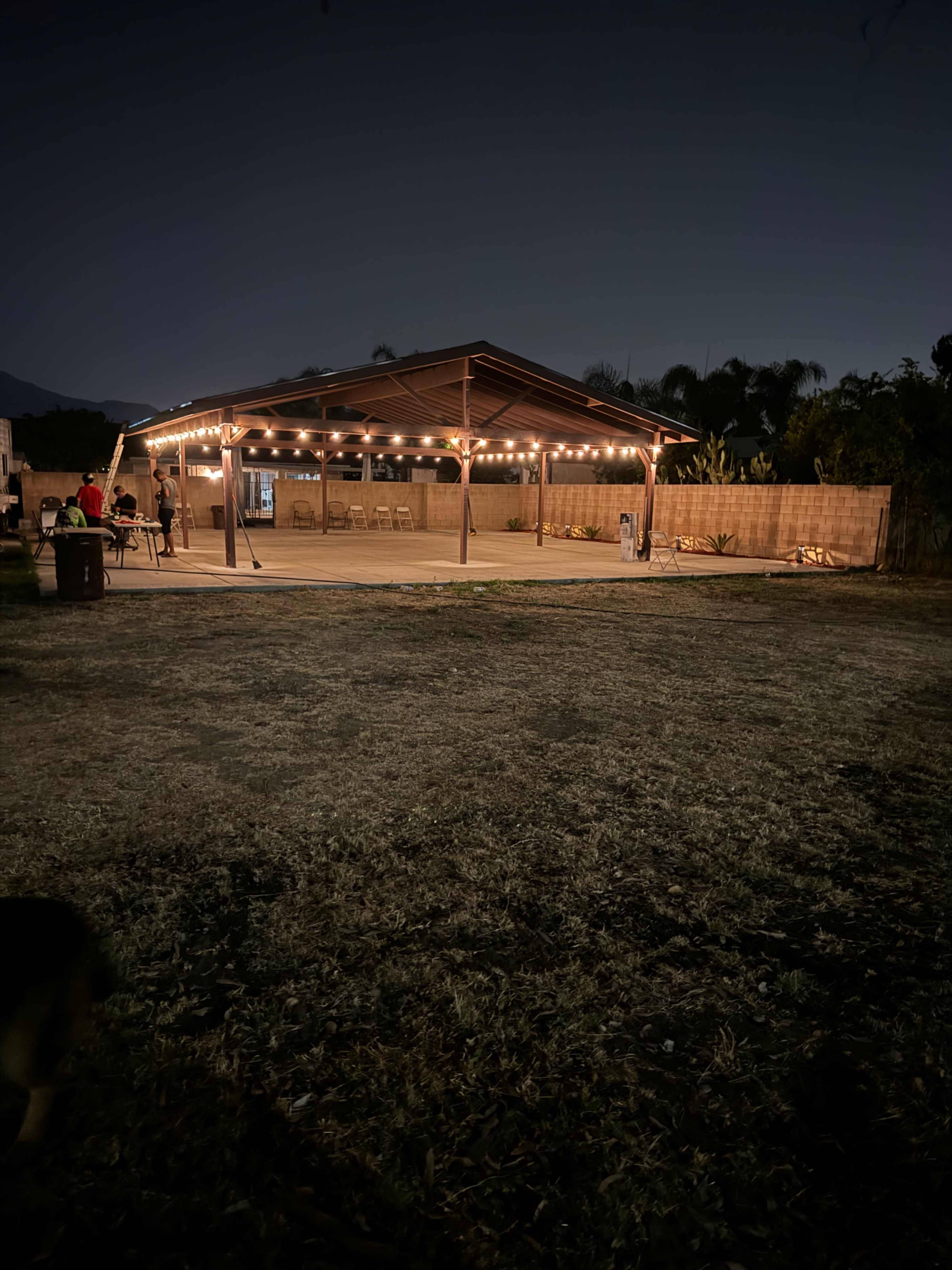 A covered outdoor venue with string lights is set up for an evening gathering, surrounded by a dark landscape.