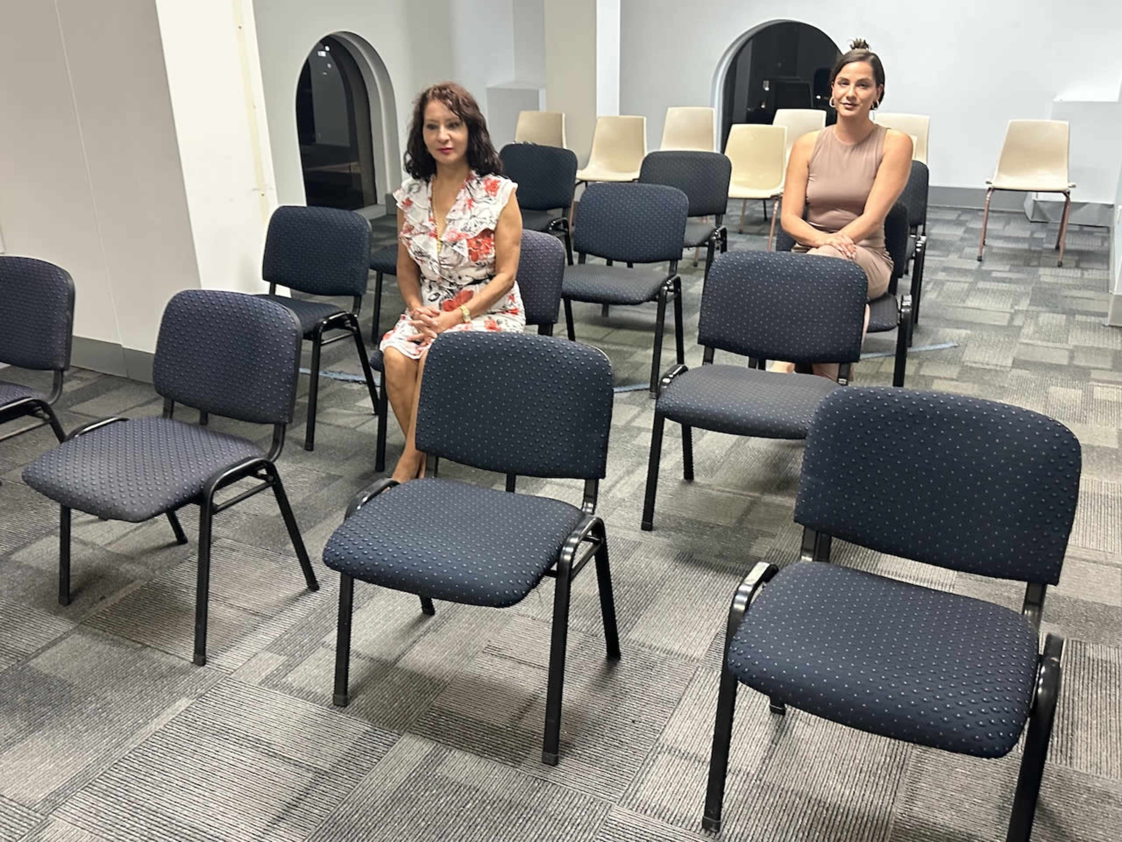 Two women sit in an empty room with rows of chairs arranged around them.