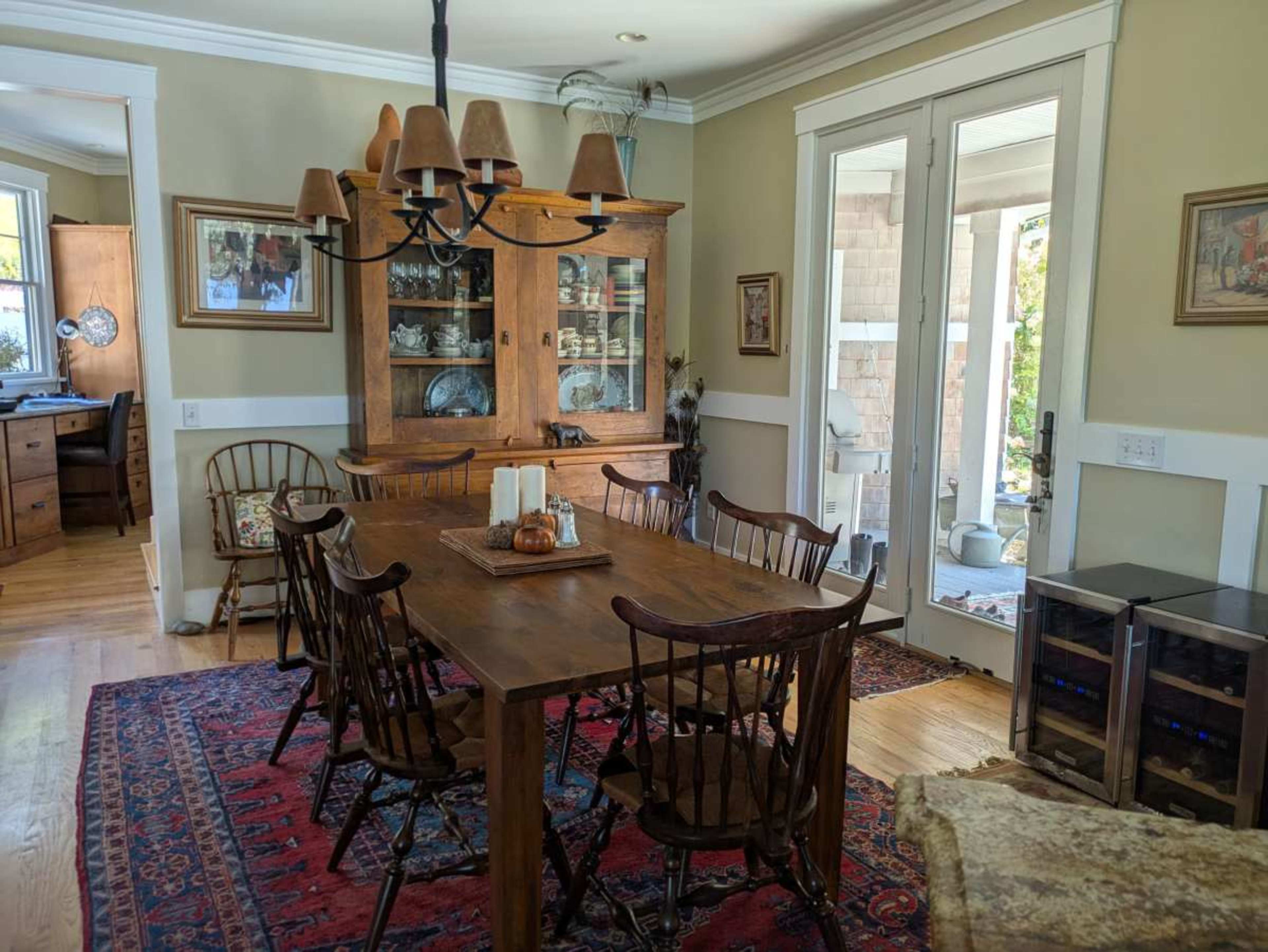 A wooden dining table surrounded by chairs sits in a well-lit room with a china cabinet, rugs, and access to a porch.