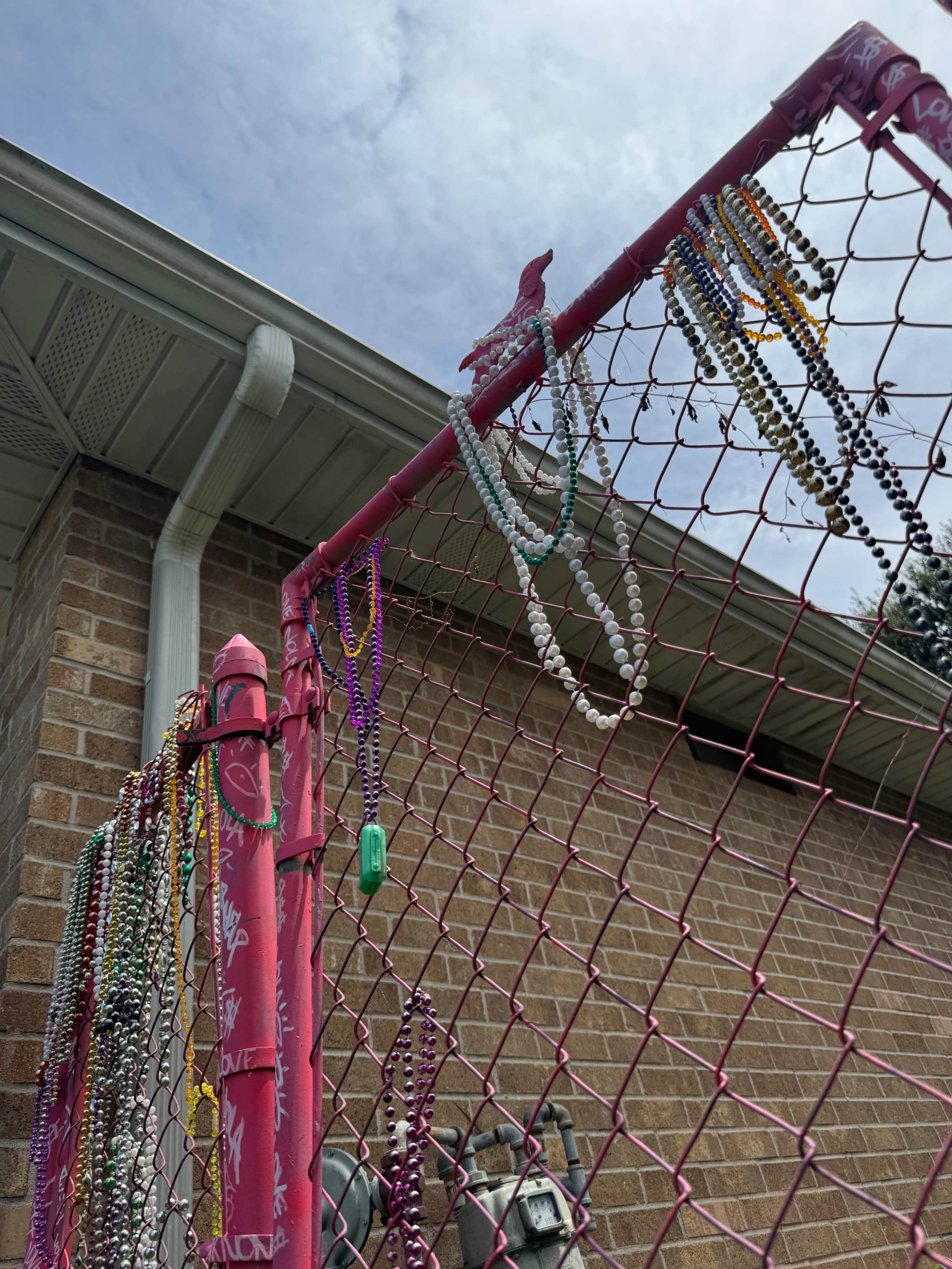 A pink chain-link fence displays colorful beads hanging from its wires against a brick wall and a partly cloudy sky.