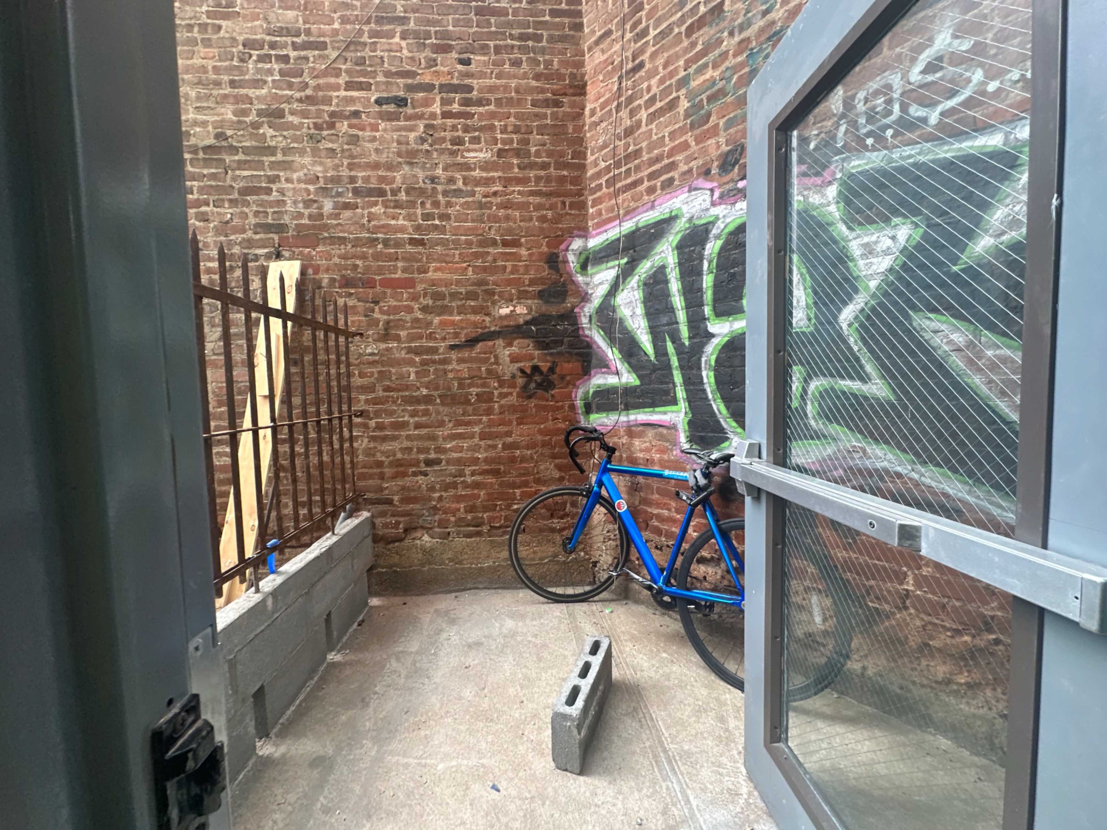 The image shows a narrow alleyway with a brick wall featuring graffiti, and a blue bicycle positioned against a concrete block.