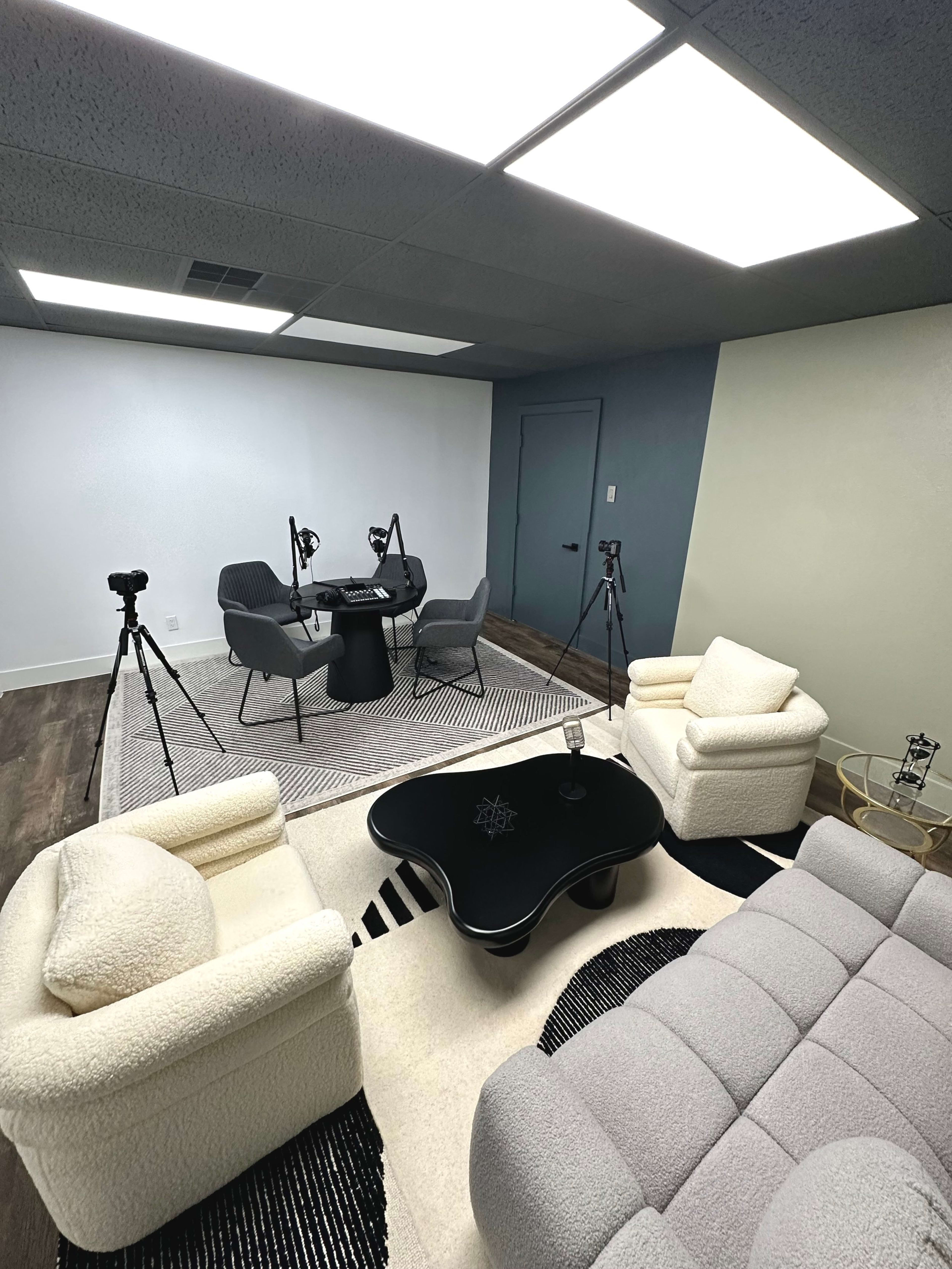 The image shows a modern, minimalistic room furnished with a white rug, a black coffee table at the center, and several cameras positioned around a round table surrounded by gray chairs.