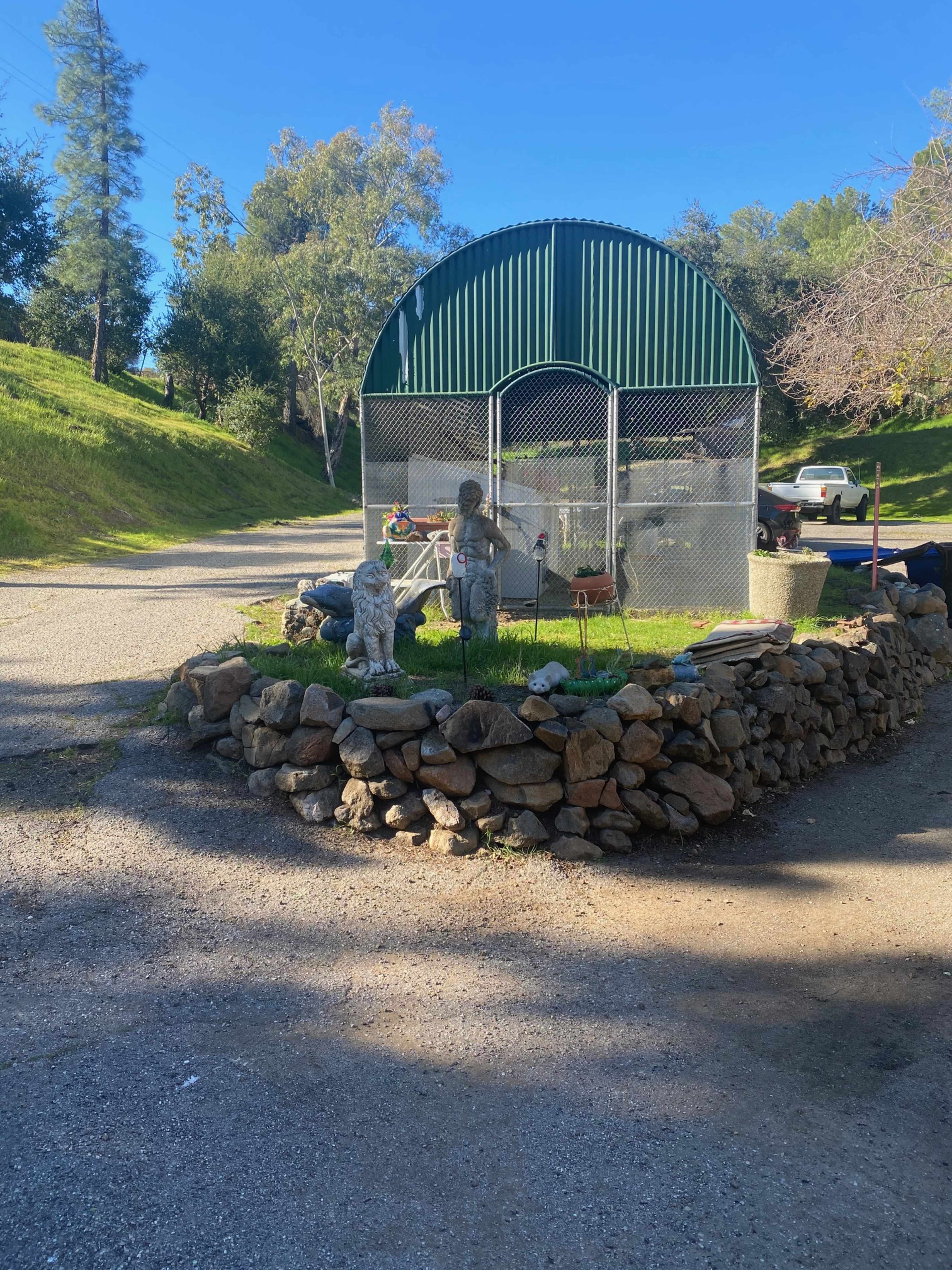 A fenced enclosure with a metal roof sits on a stone-bordered area, surrounded by grass and trees.