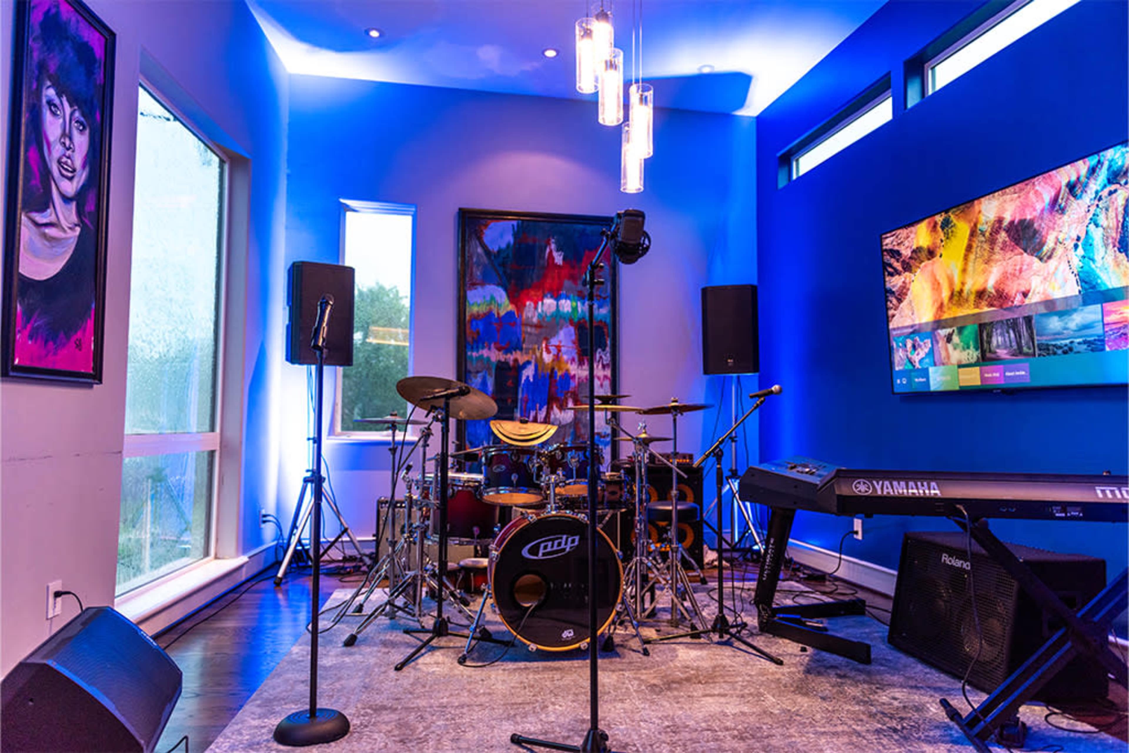 The image shows a music rehearsal space featuring a drum set, keyboard, and various audio equipment against a backdrop of colorful artwork and ambient lighting.