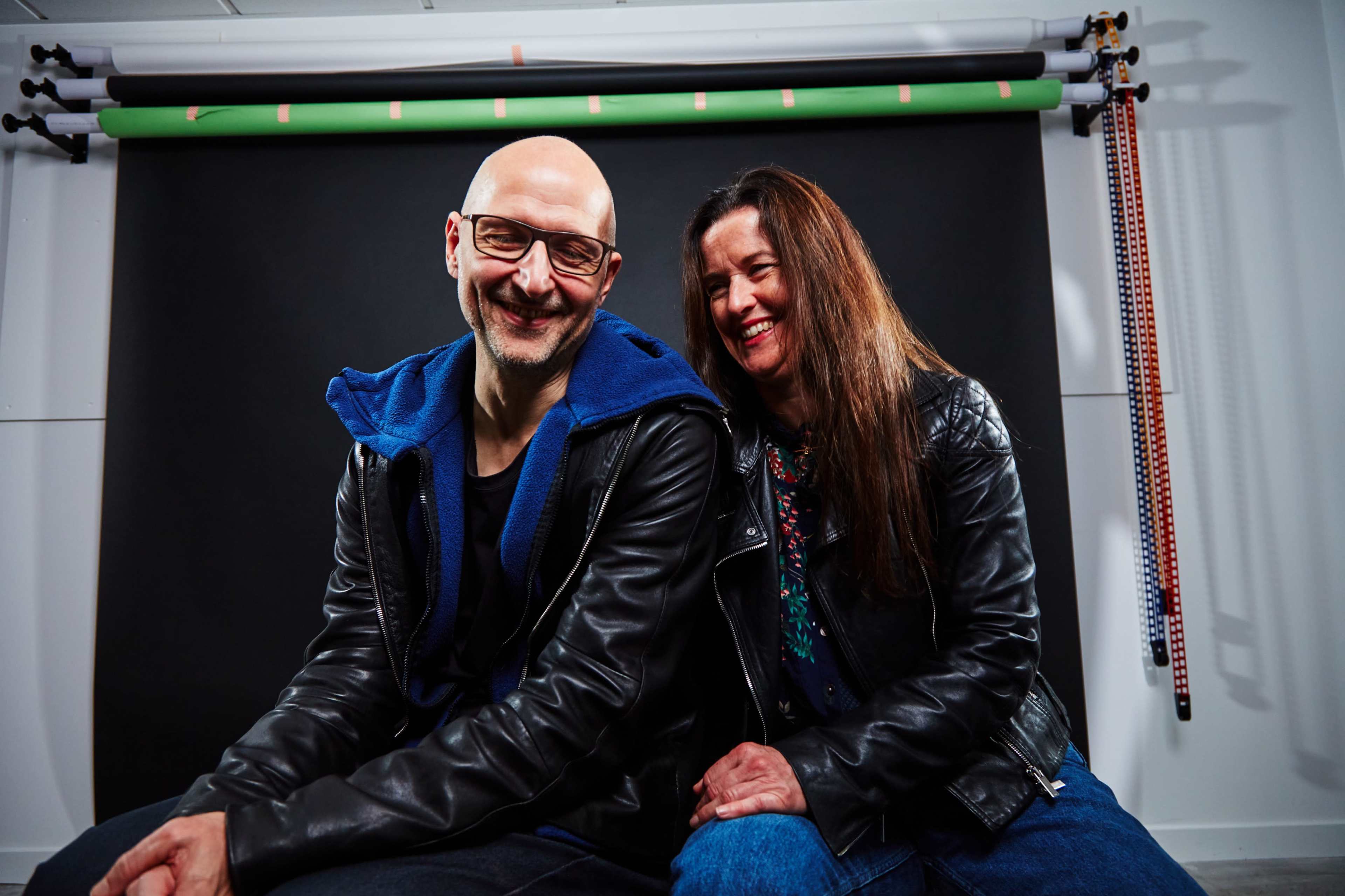 A man and a woman sit closely together on a studio set with a black backdrop and colorful equipment visible in the background.
