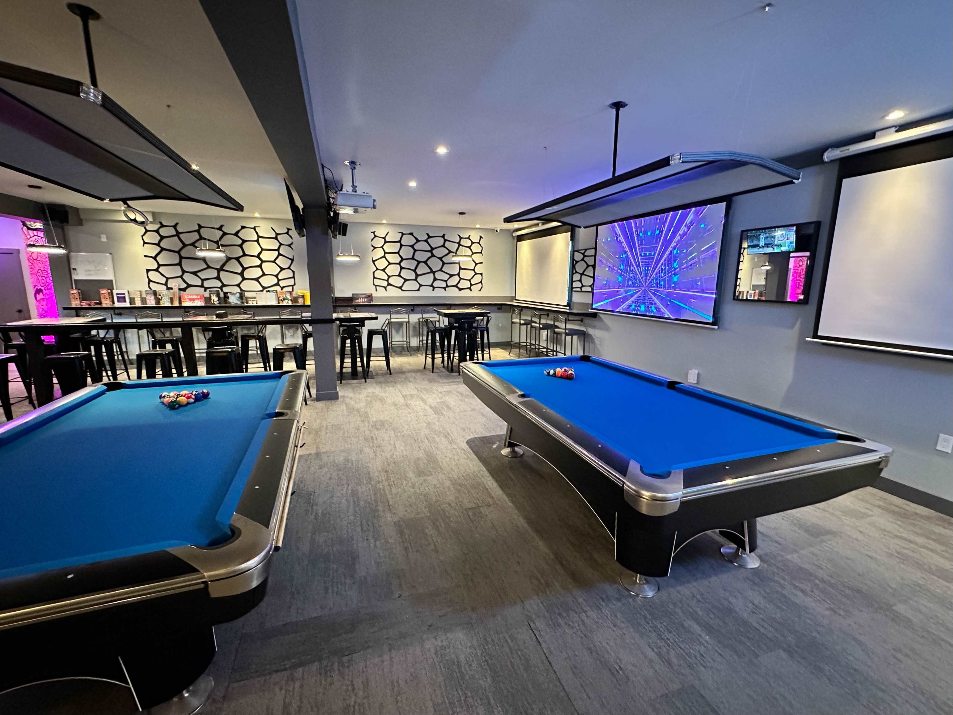 A room with two pool tables, neon lighting, and a projector screen displaying colorful graphics on the wall.