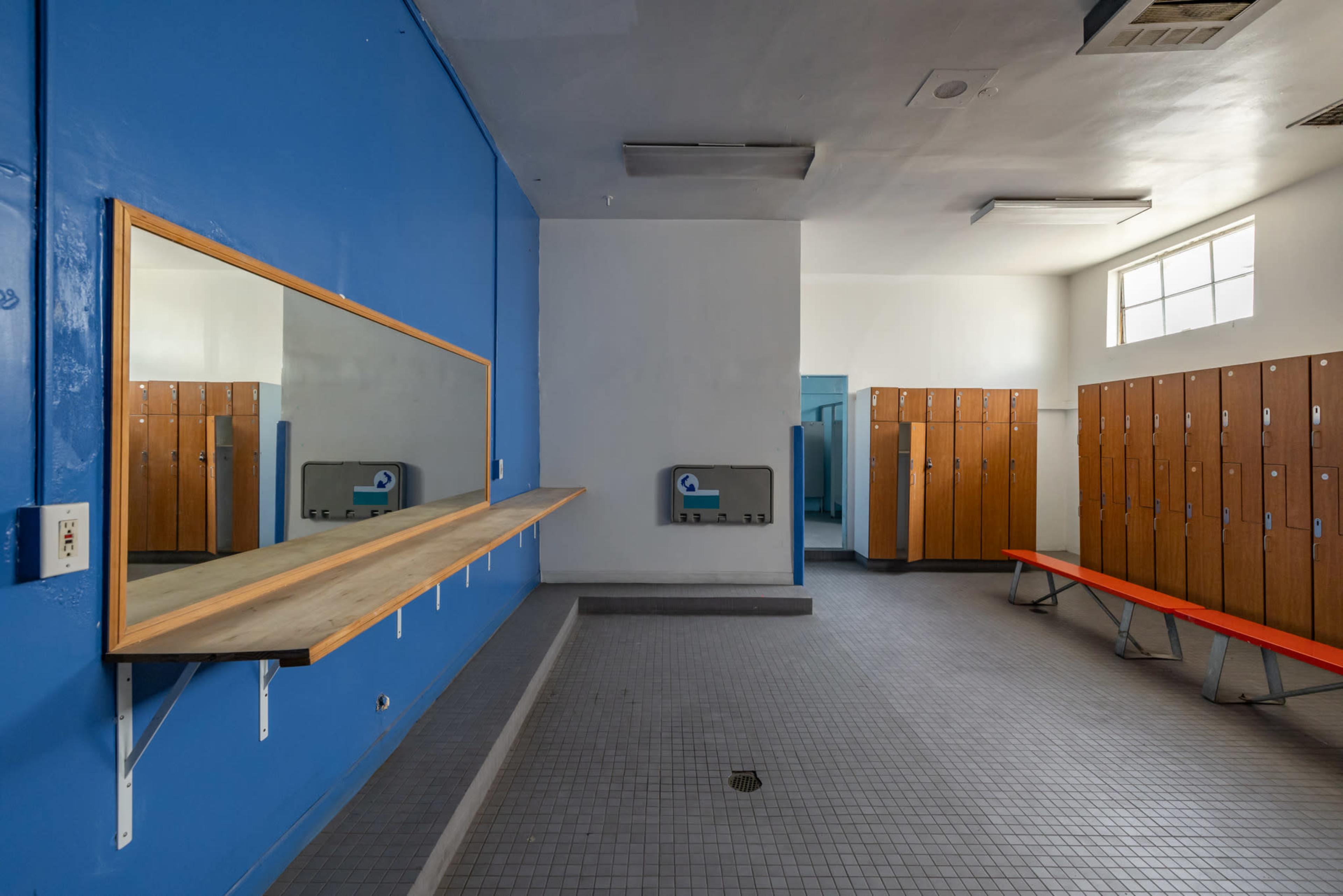 An empty locker room features blue walls, a wooden bench with a mirror, and rows of wooden lockers.