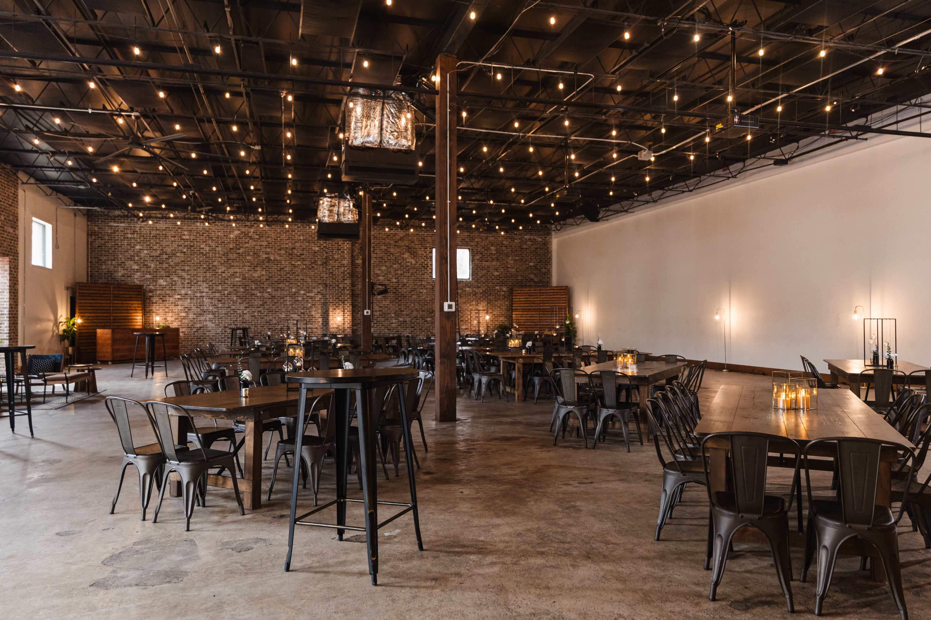 The image shows a spacious event venue with wooden tables and metal chairs, adorned with string lights and a rustic brick wall.