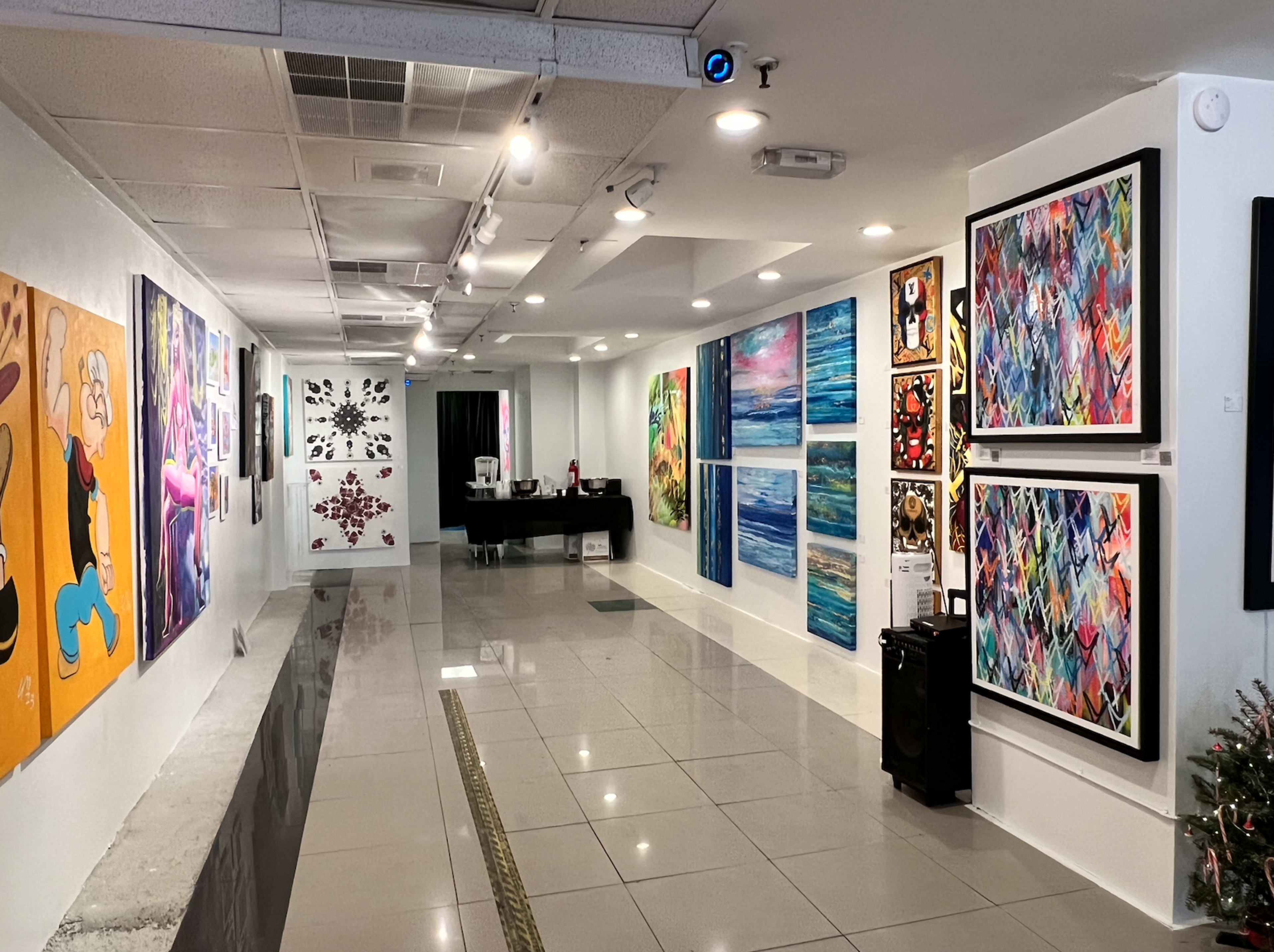 An art gallery with colorful paintings displayed along white walls and a reception area at the end of a tiled hallway.