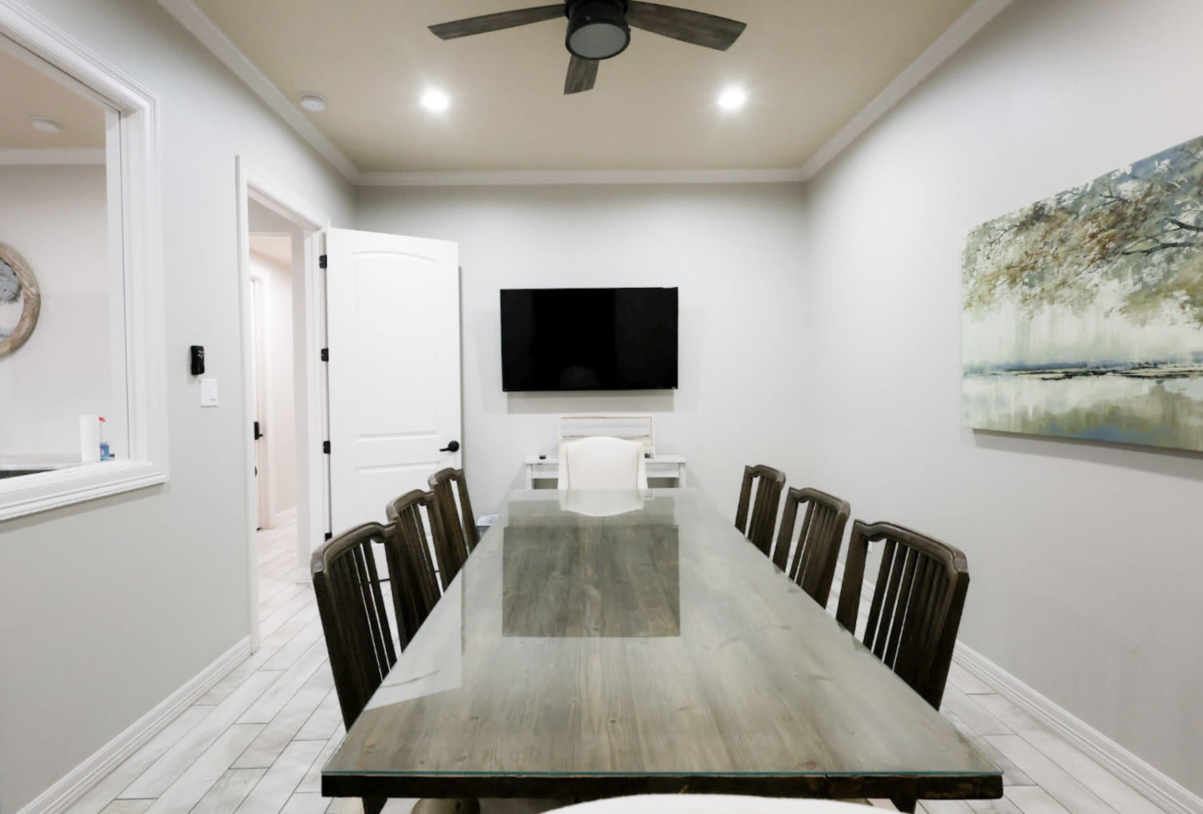 A long wooden dining table is set in a light-colored room with a large wall-mounted TV and a painting on the adjacent wall.