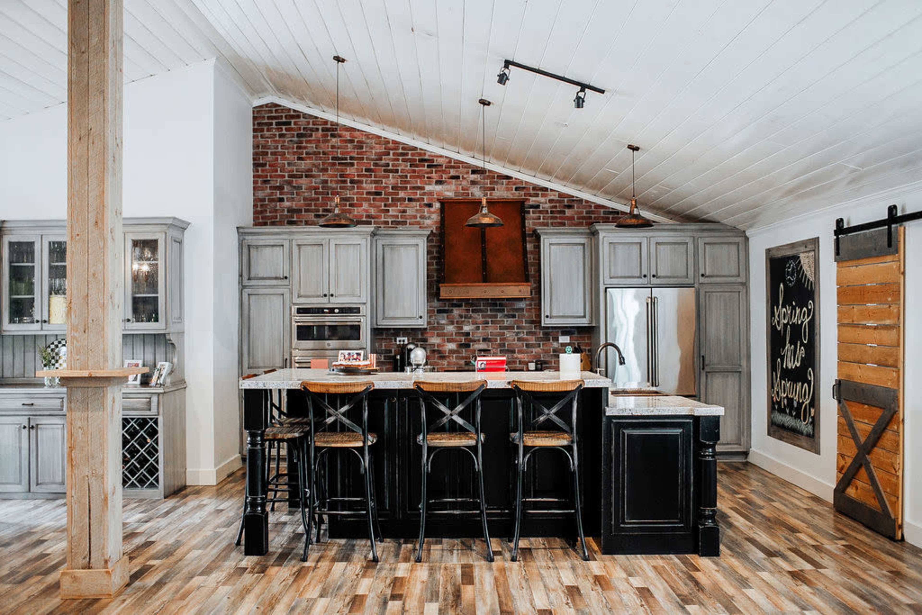 A modern kitchen featuring gray cabinets, a brick accent wall, and a large island with bar stools.