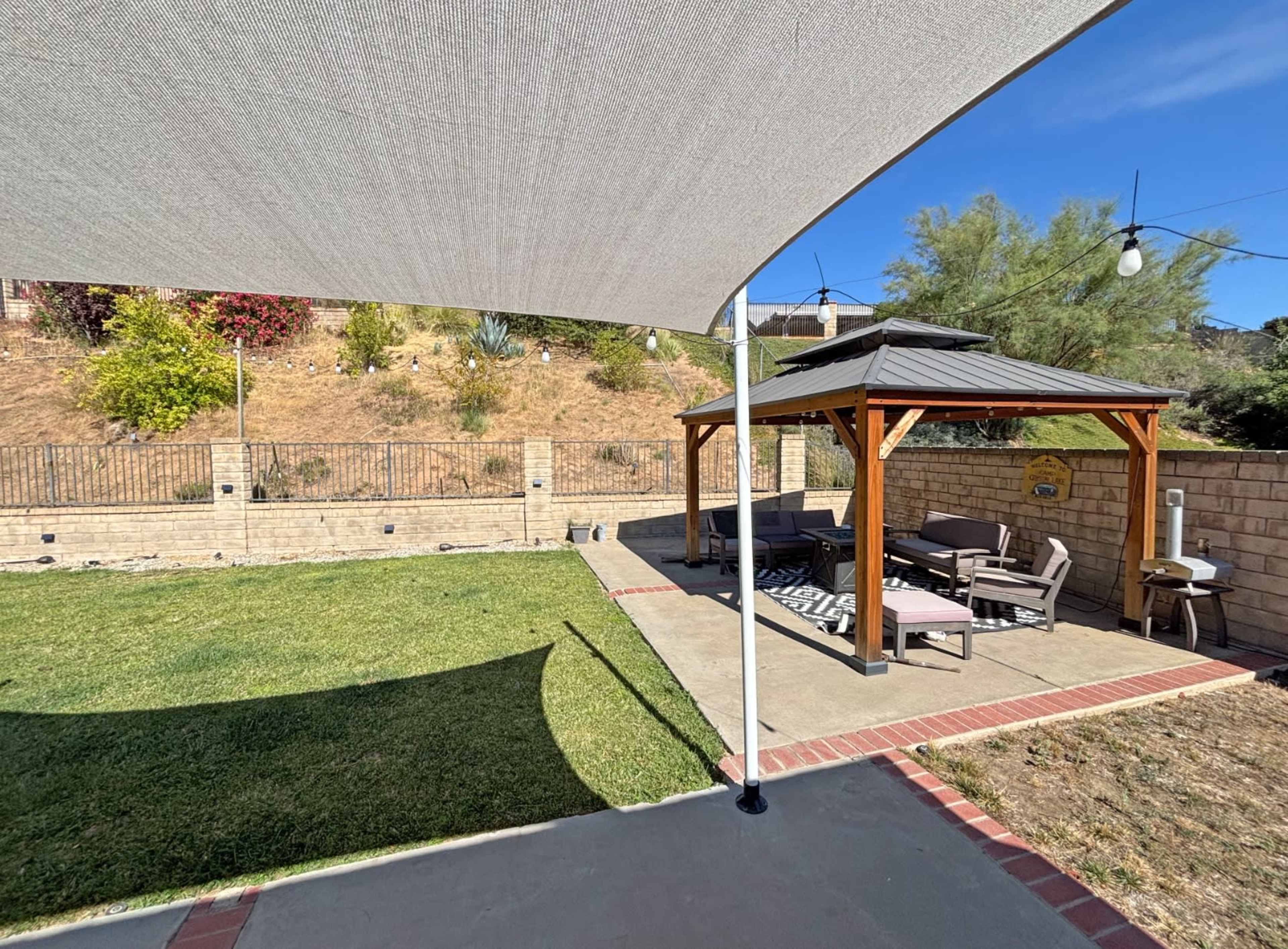 The image shows a backyard with a shaded wooden gazebo, a grassy area, and decorative string lights in the background.