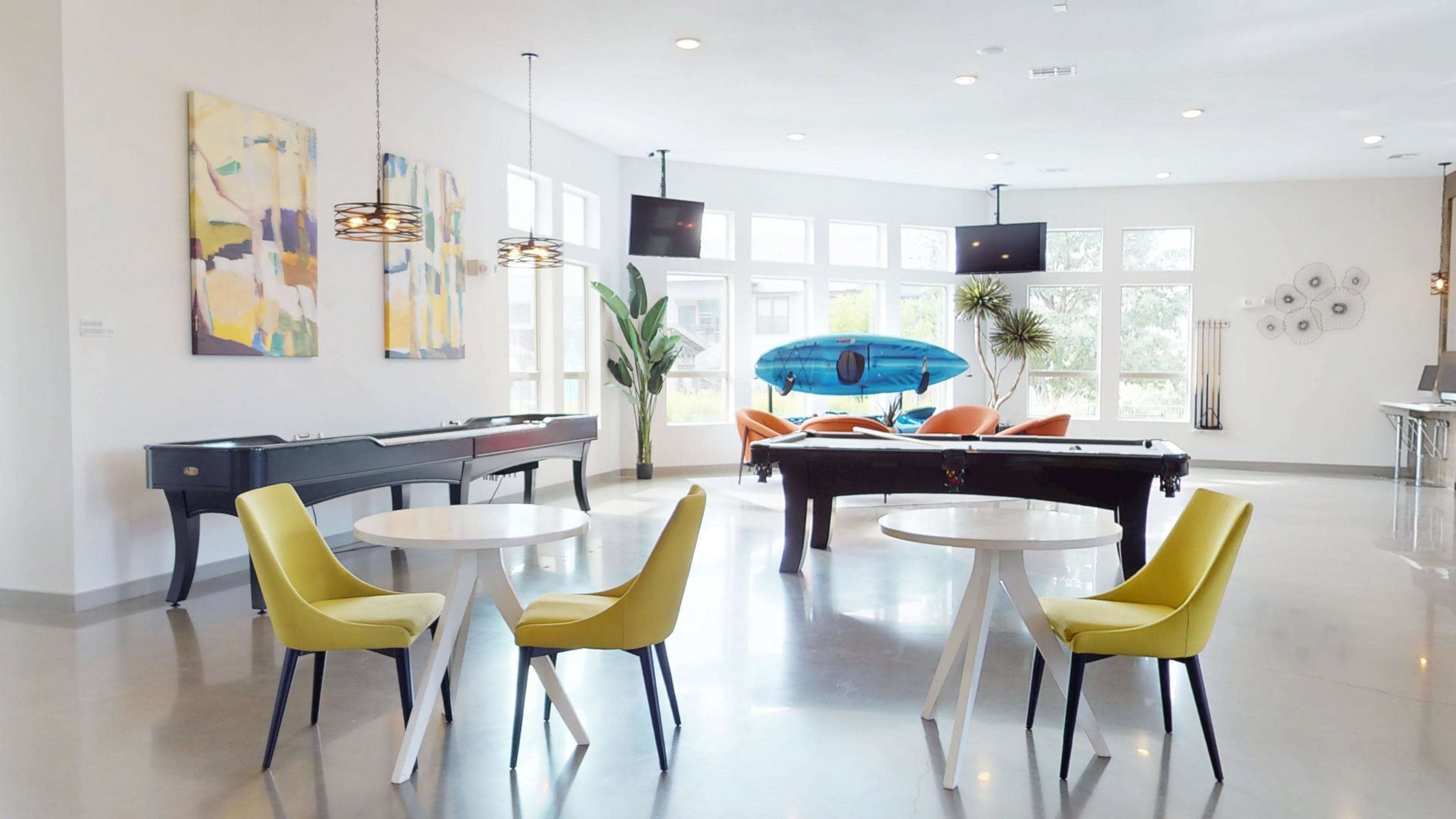 The image shows a spacious communal area featuring a pool table, a kayak on display, modern seating arrangements, and large windows allowing abundant natural light.