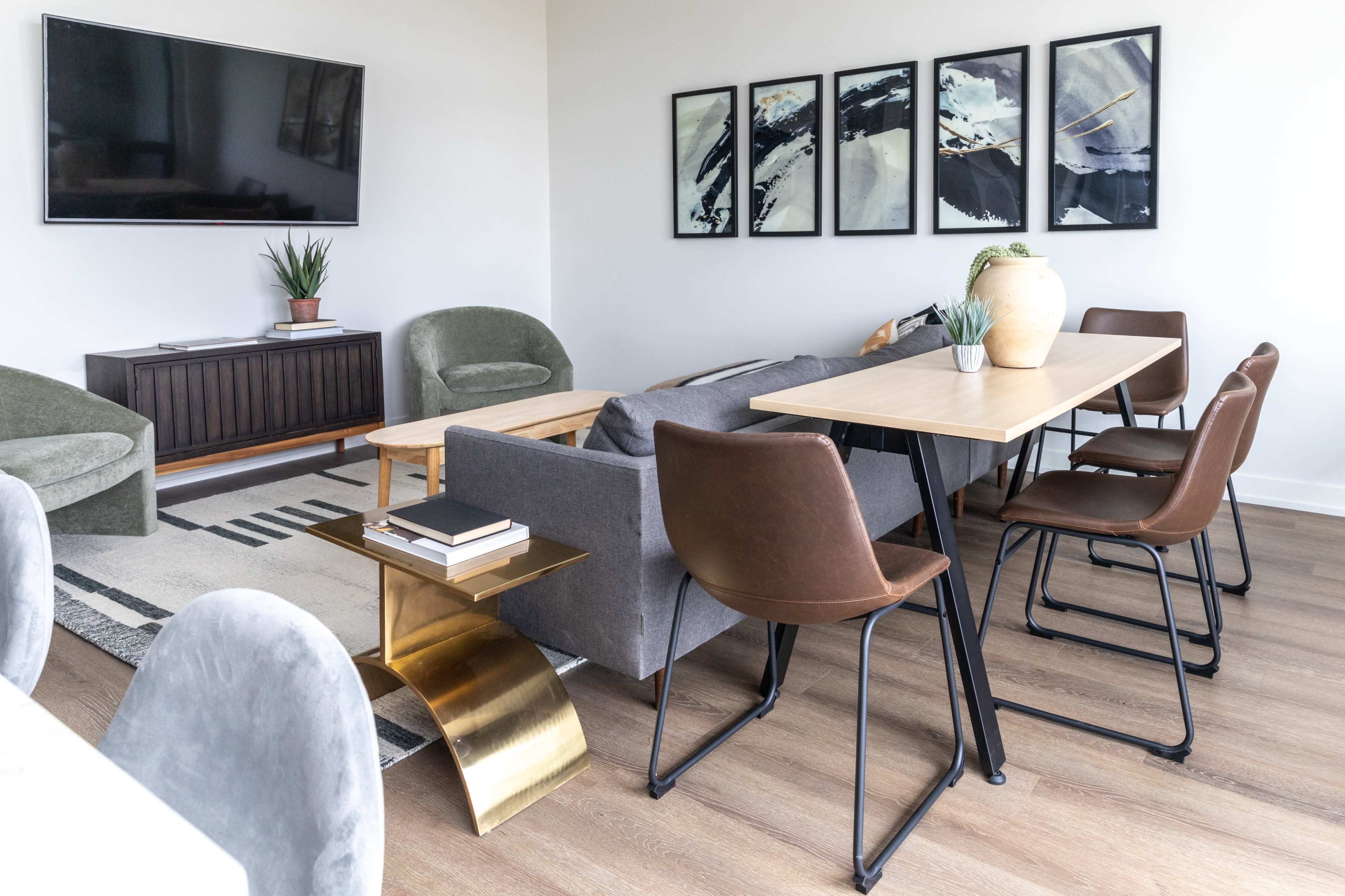 A modern living area featuring a wooden coffee table, six dining chairs around a table, two green armchairs, and framed artwork on the wall.