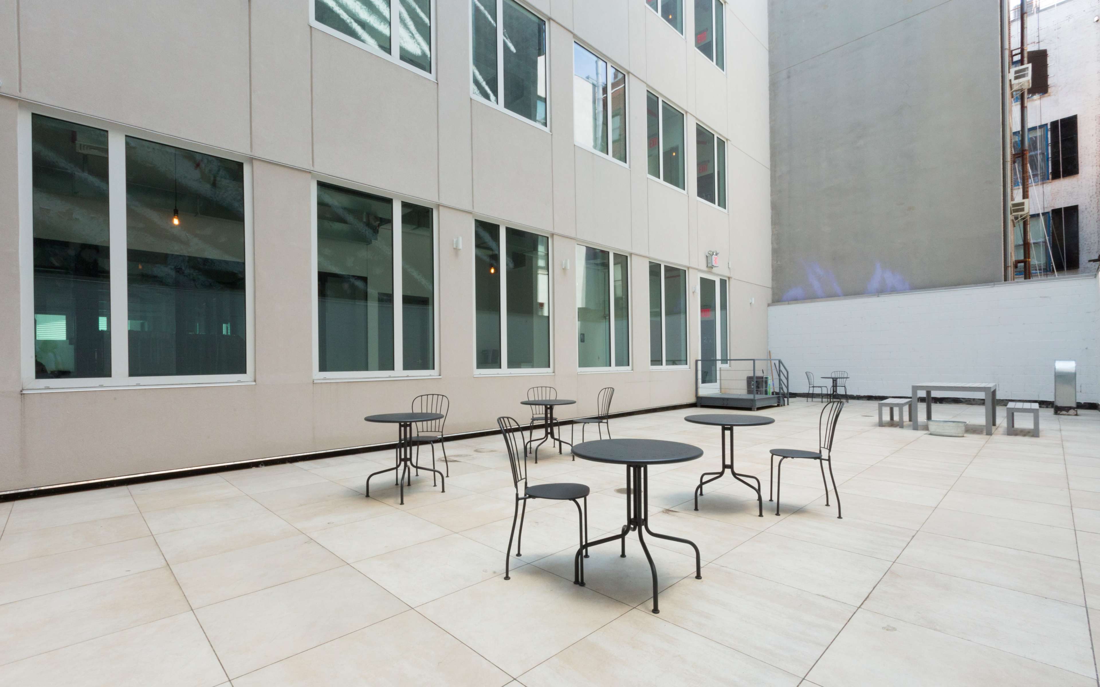 An outdoor courtyard with several small tables and chairs situated on a tiled surface, surrounded by tall building walls.