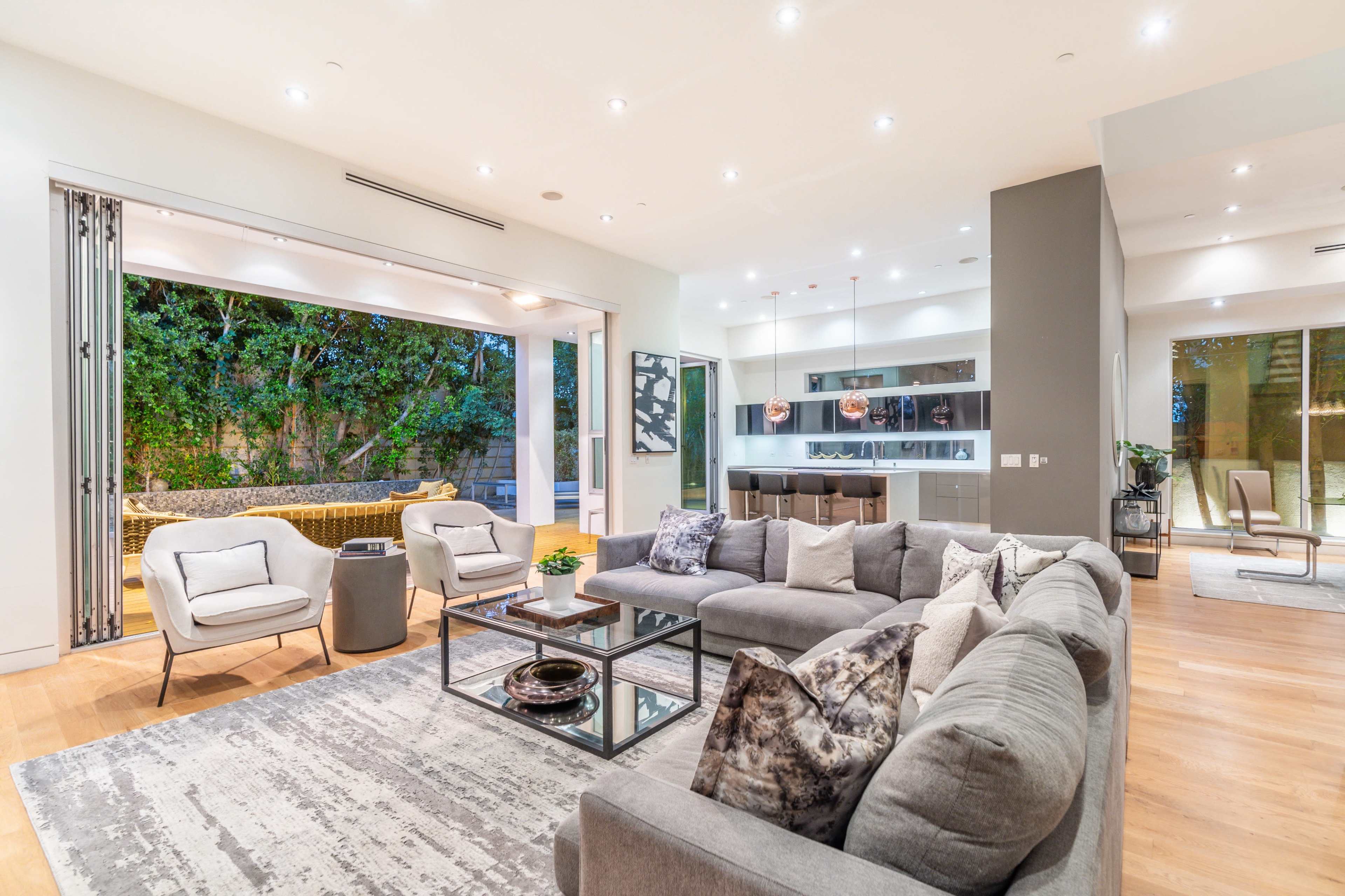 A modern living room with a large sectional sofa, a glass coffee table, and an open view into an outdoor area framed by sliding glass doors.