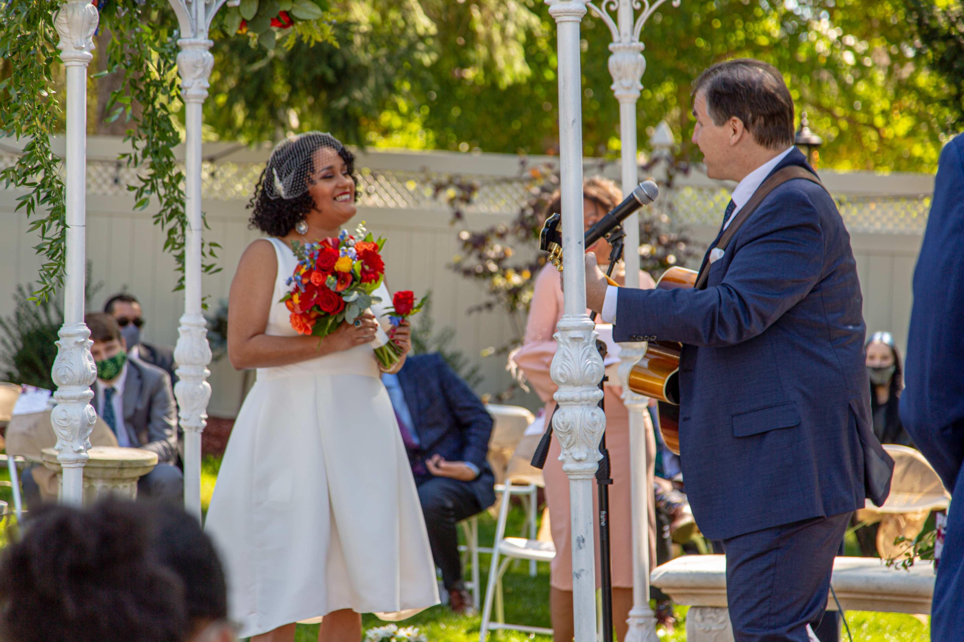 A bride in a white dress stands under a wedding arch, holding a bouquet, while a man in a blue suit plays guitar and sings.