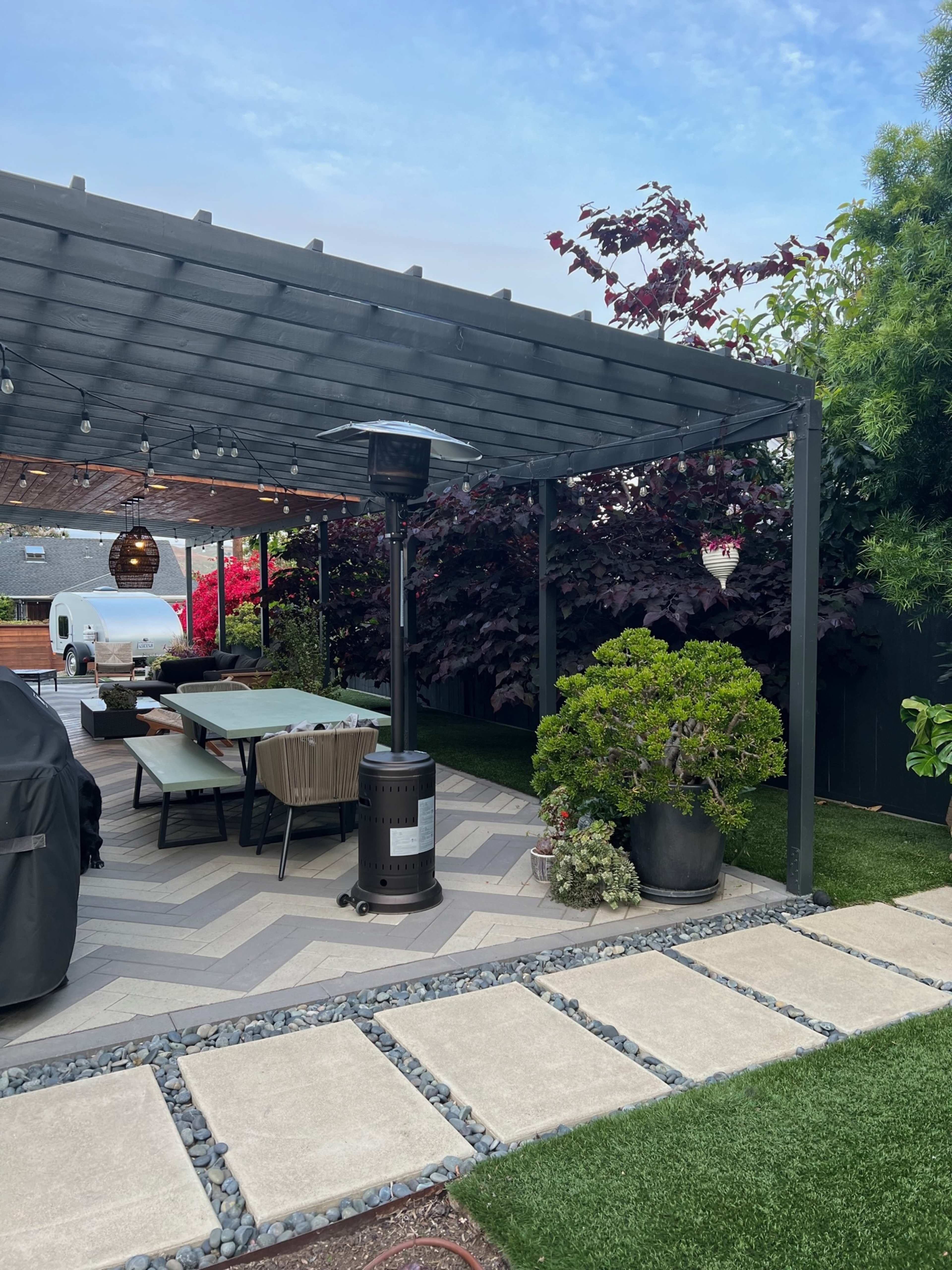 A covered patio area features a dining table and heating lamp, with a manicured garden and decorative plants alongside a stone pathway.