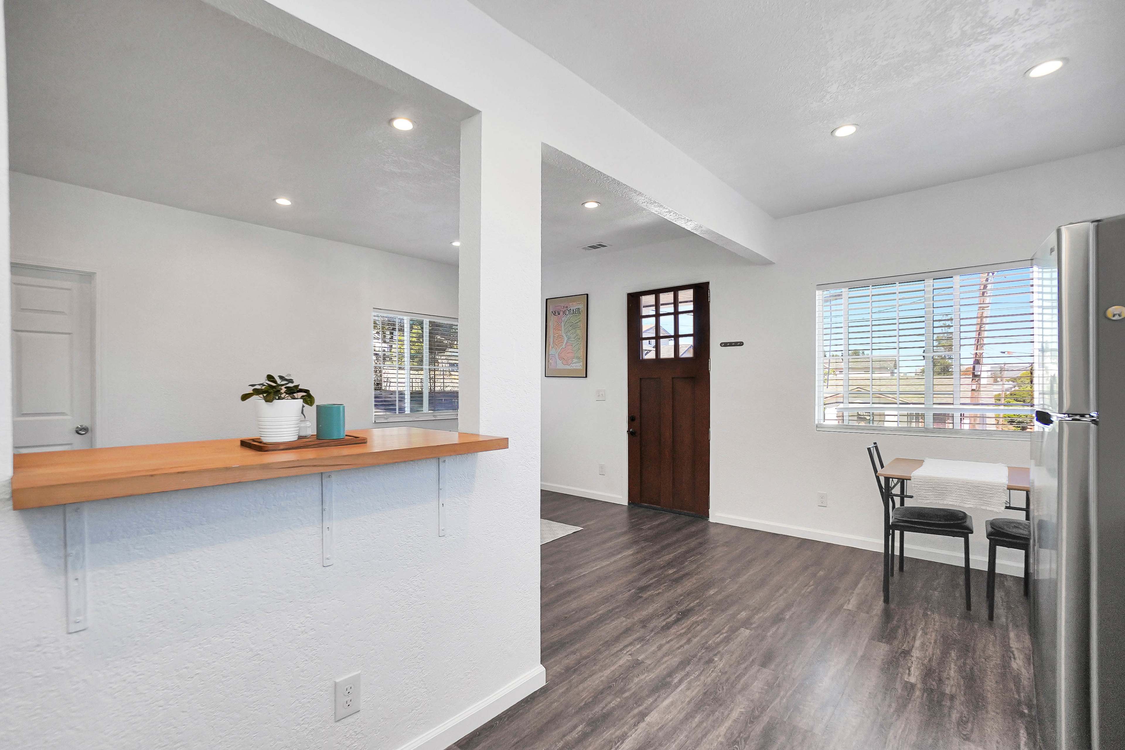 A bright, modern kitchen area features a dining table by a window, with a doorway leading to a living space and a dark wood front door.