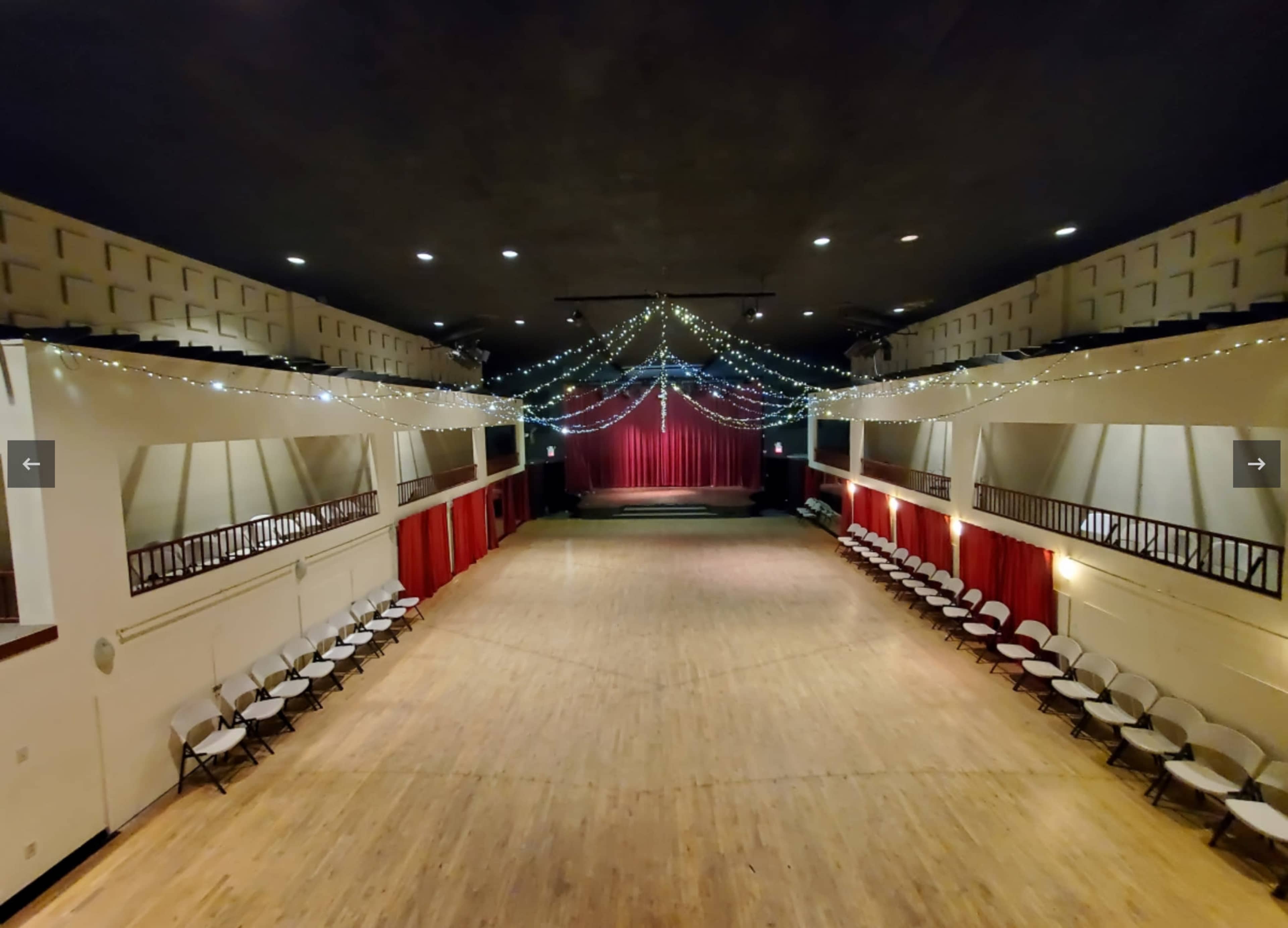 The image shows a spacious auditorium with rows of white chairs arranged on either side, a wooden floor, and decorative string lights draping from the ceiling above a red curtain stage.