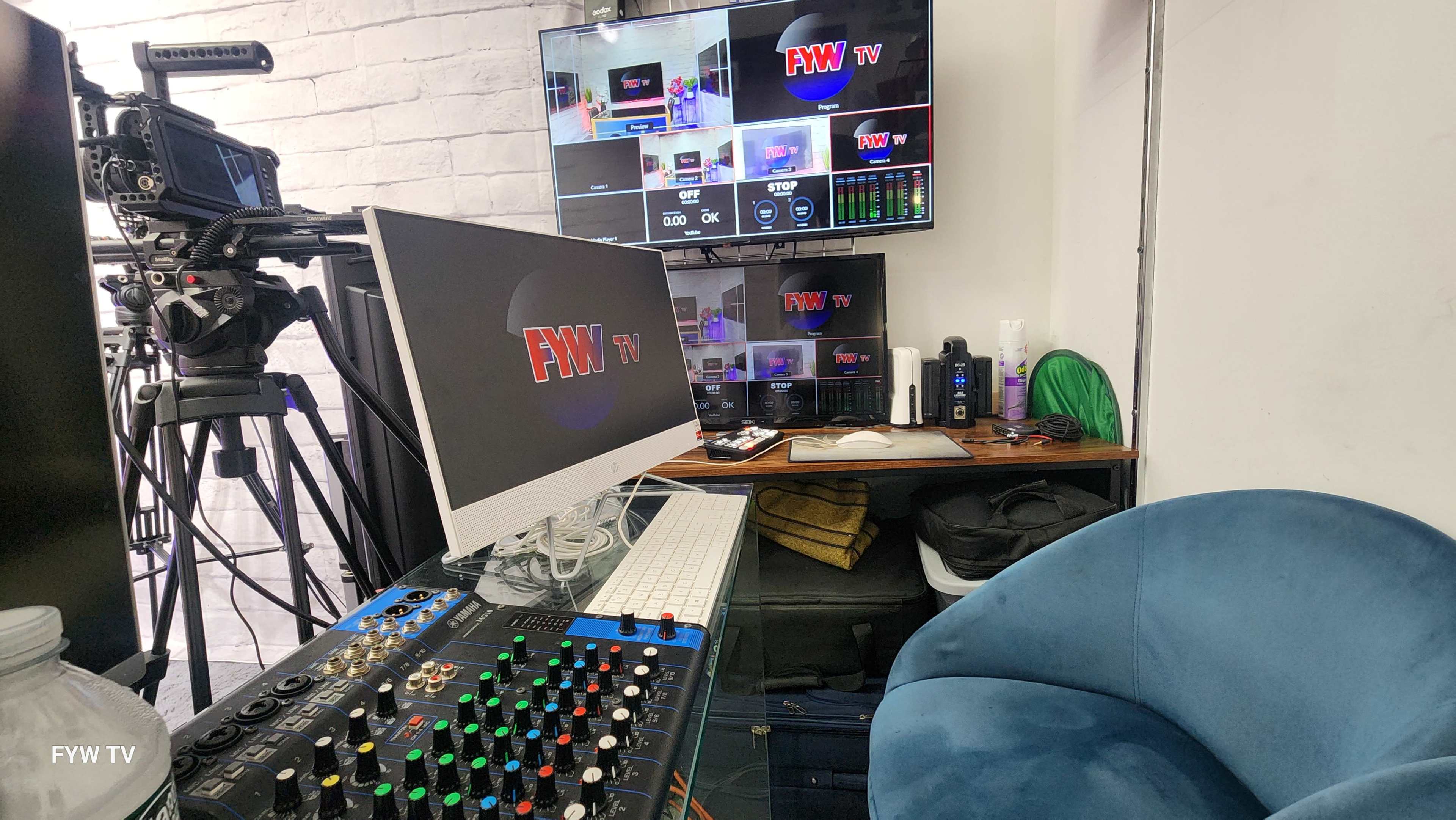 A television production setup features multiple monitors displaying live feeds, a computer with audio equipment, and a blue chair in a studio environment.