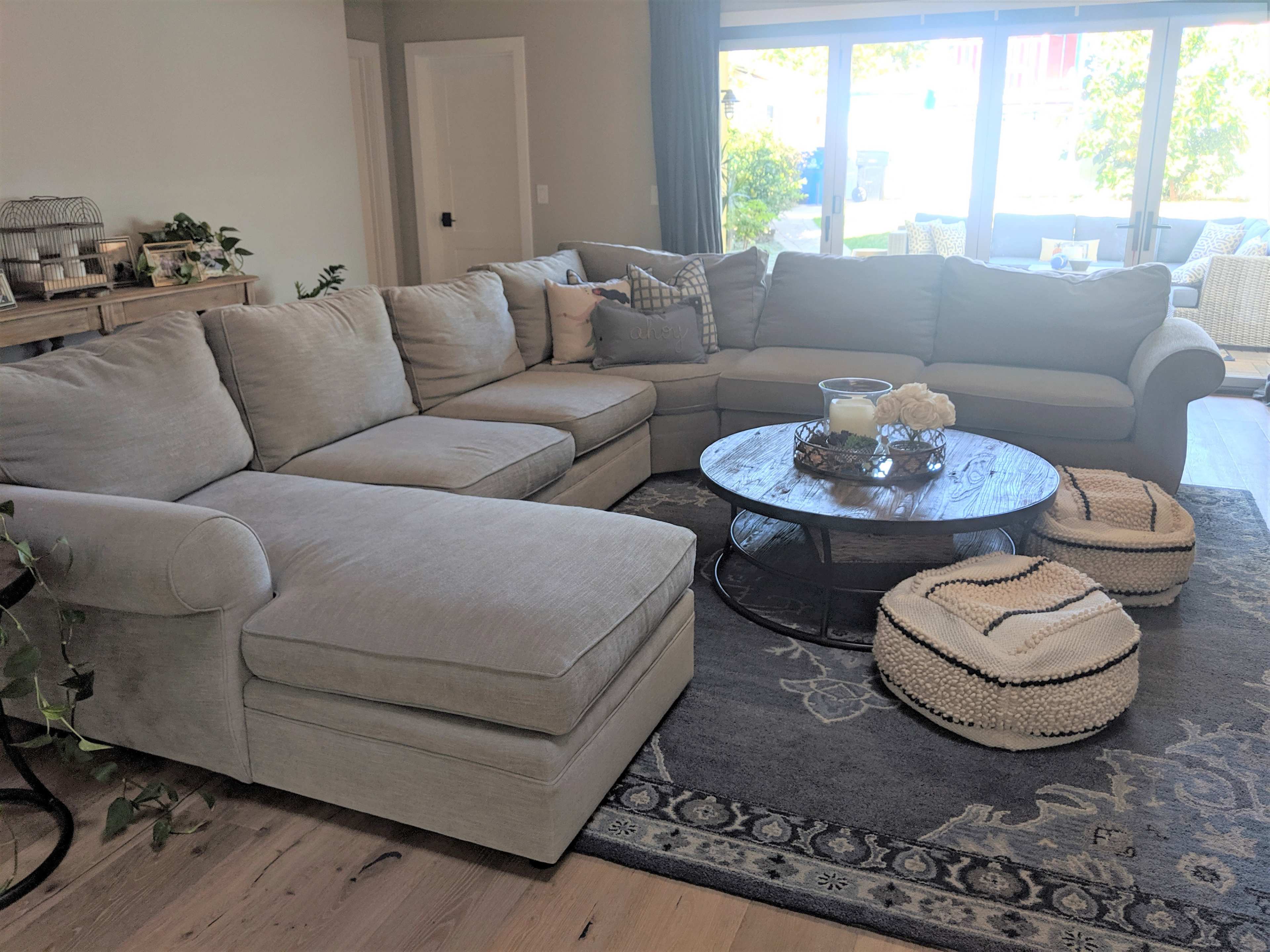 A large, light gray sectional sofa is arranged around a round wooden coffee table on a decorative area rug in a bright, open living space with large windows.