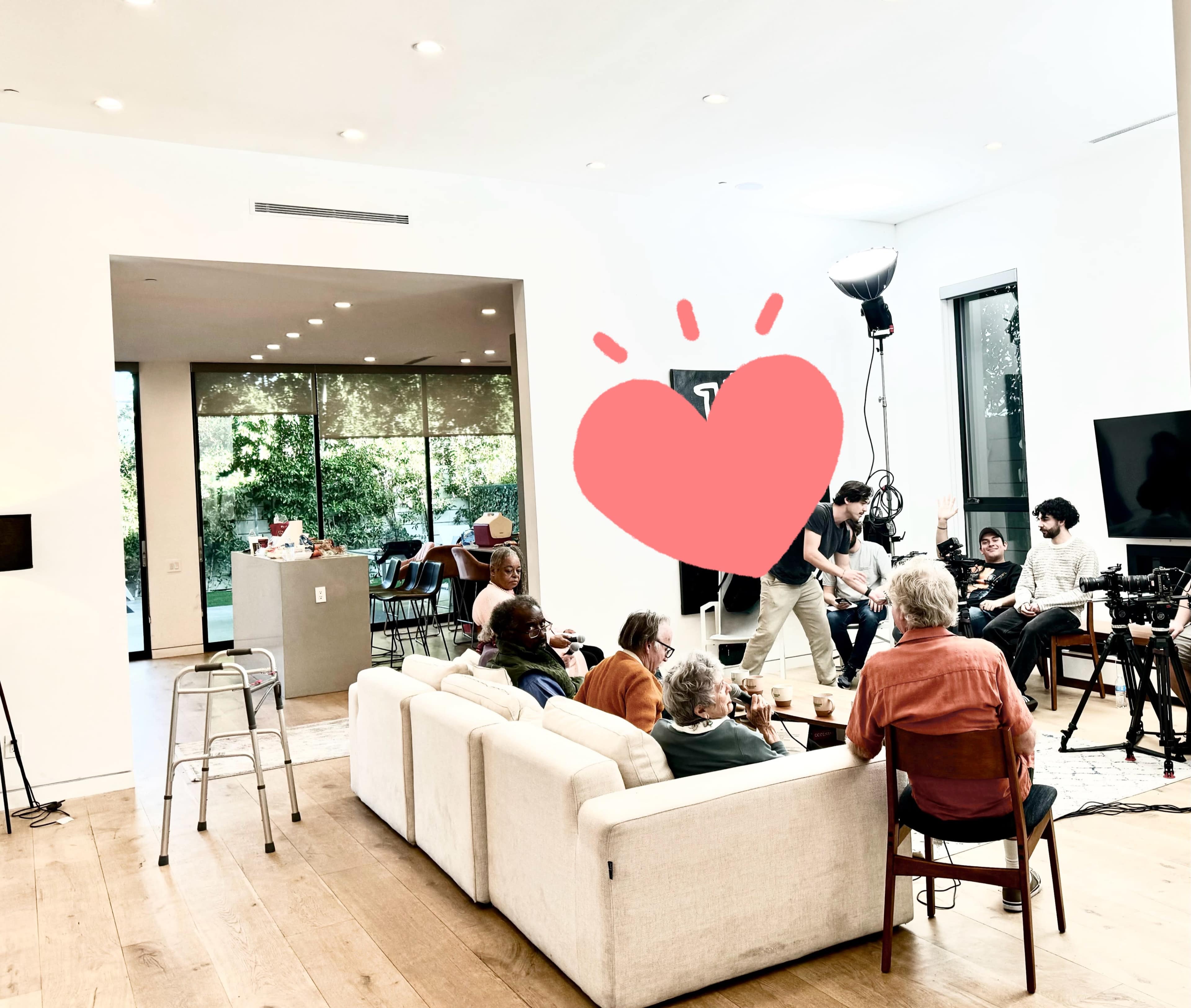 A group of people is seated on a couch in a well-lit room, while a few individuals set up filming equipment nearby.