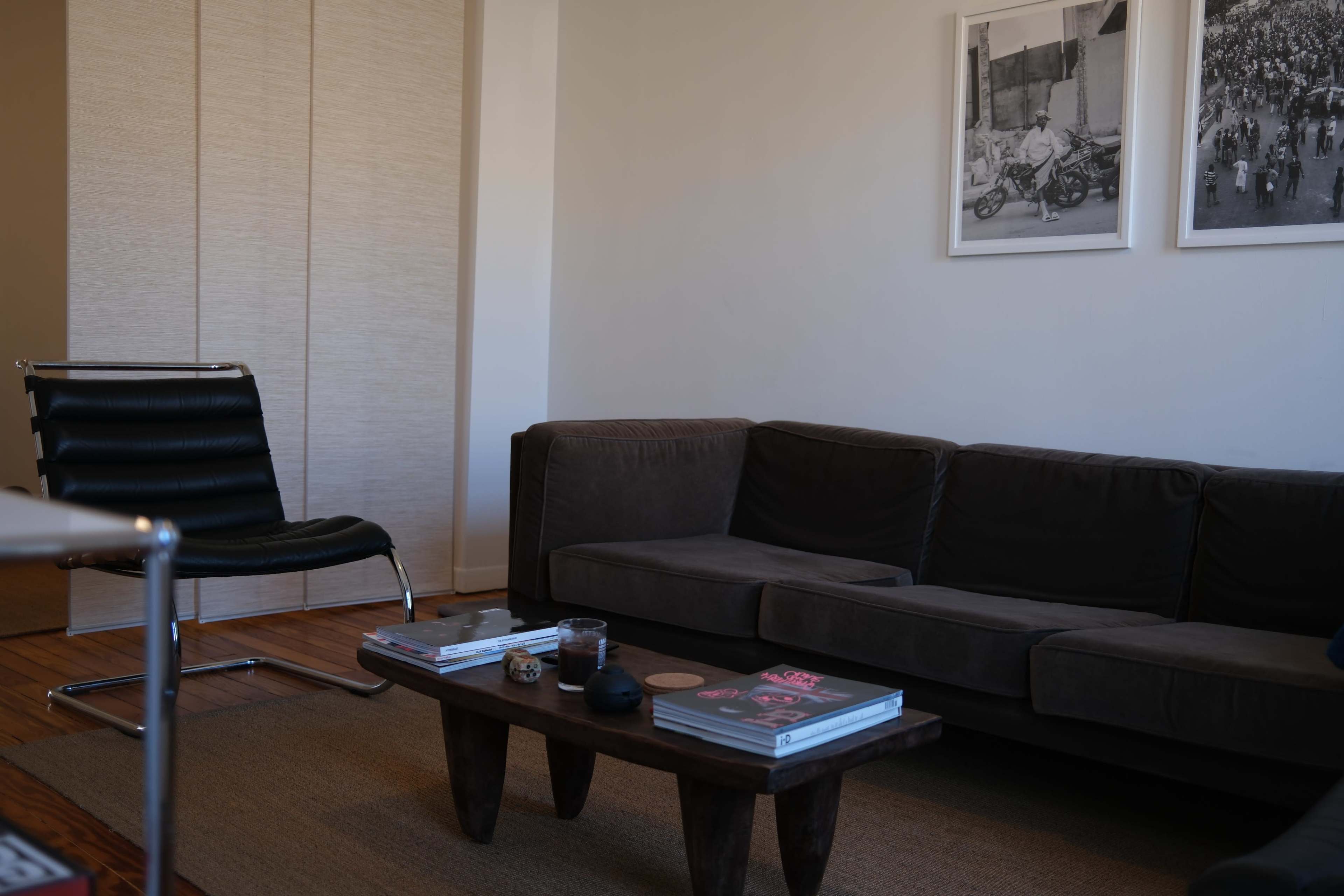 A living room features a dark sofa, a wooden coffee table with magazines, a black chair, and two framed photographs on the wall.