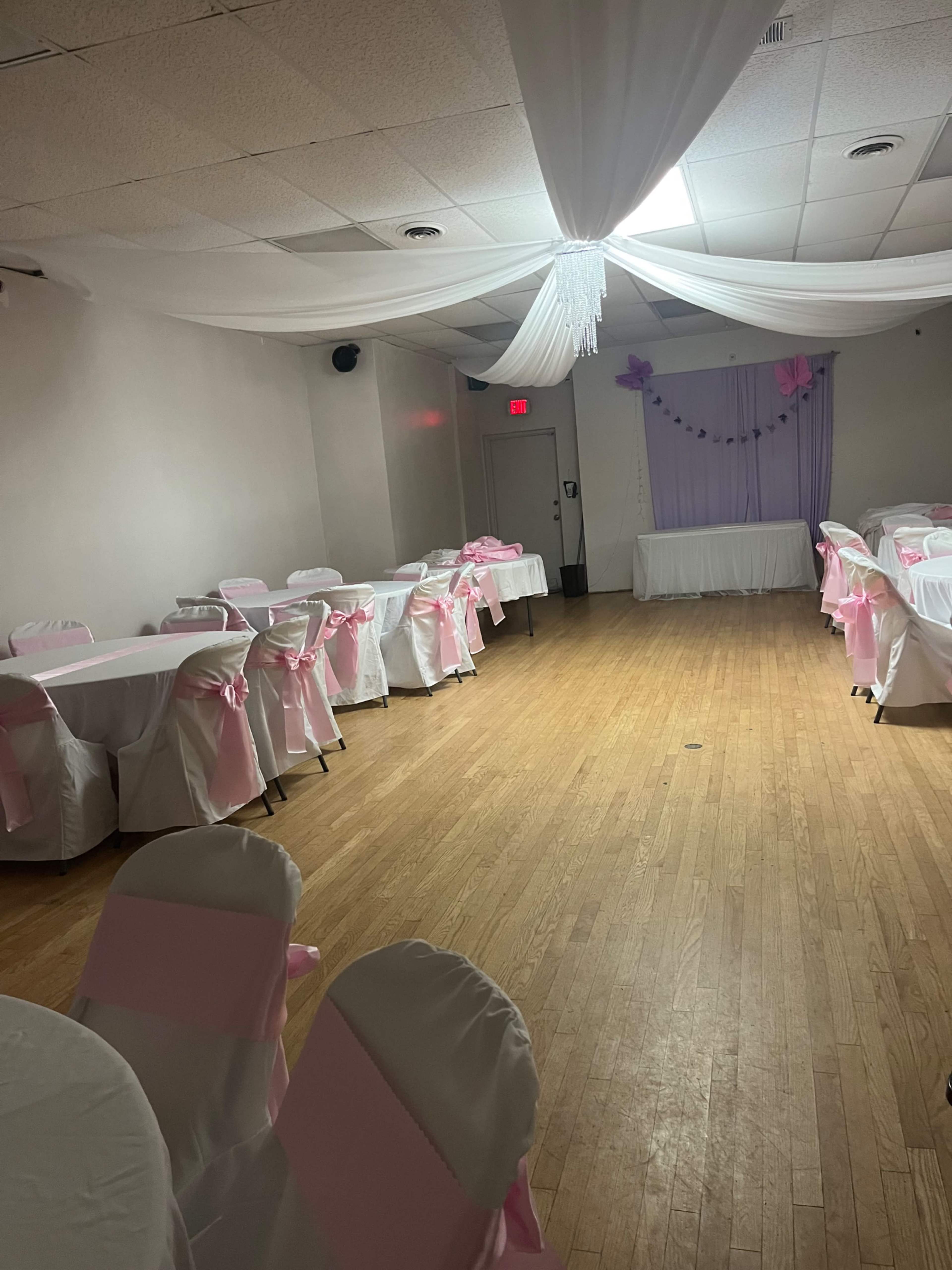 A spacious banquet hall features white tables and chairs adorned with pink sashes, with decorations hanging above and a backdrop at the front.