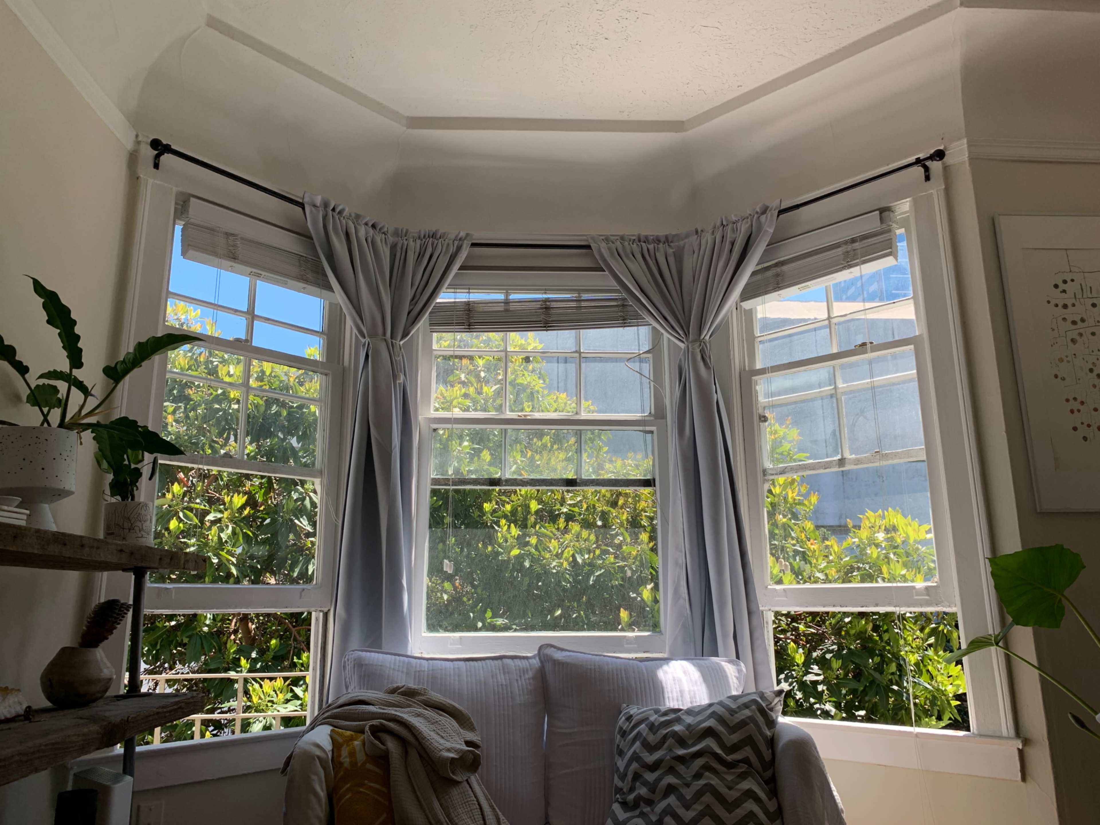A bright, airy corner of a room with large windows draped with light curtains, overlooking greenery outside.