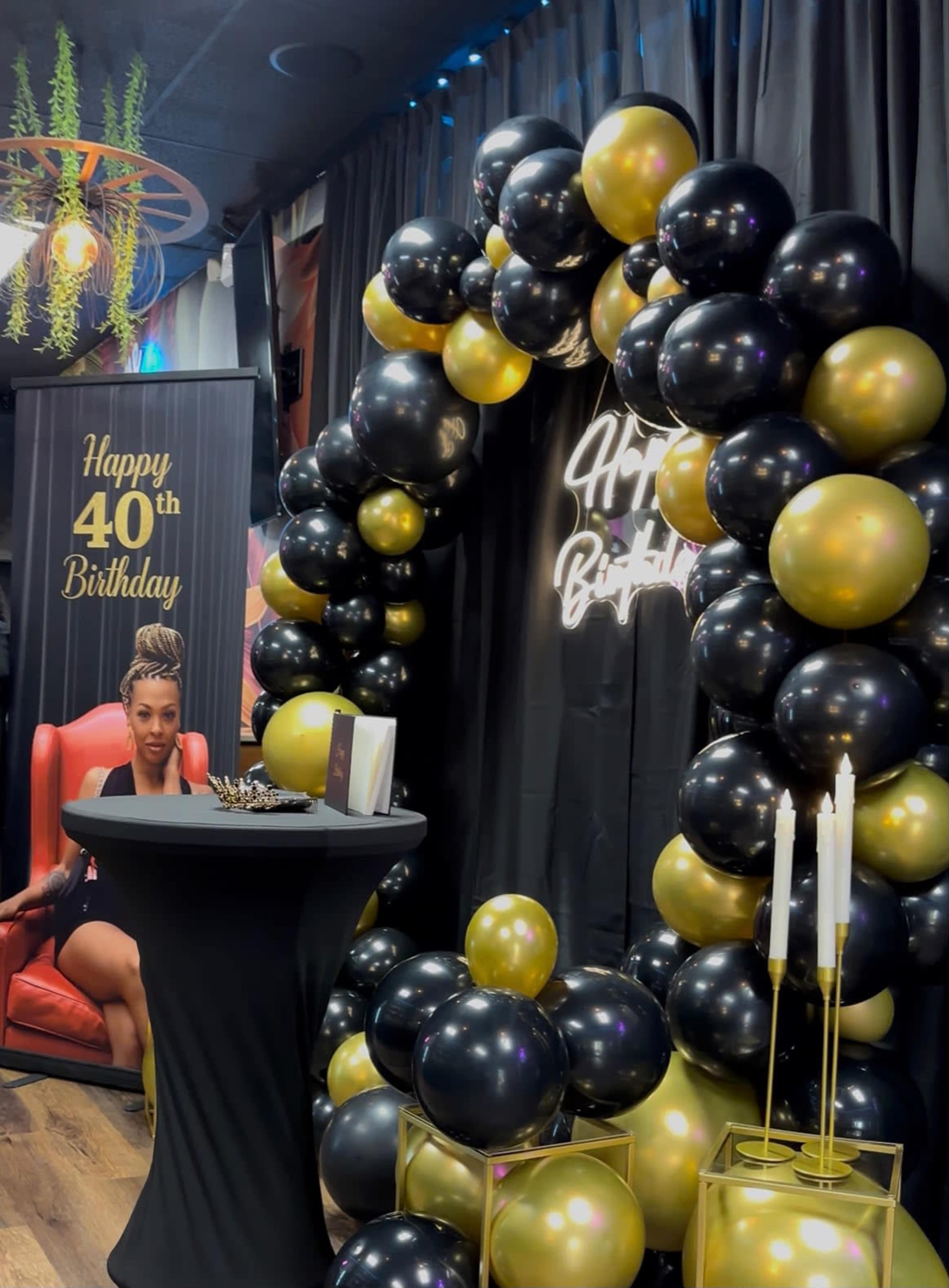 The image features a decorated party scene with a black and gold balloon arch, a birthday greeting sign, and a seated woman next to a table with candles.