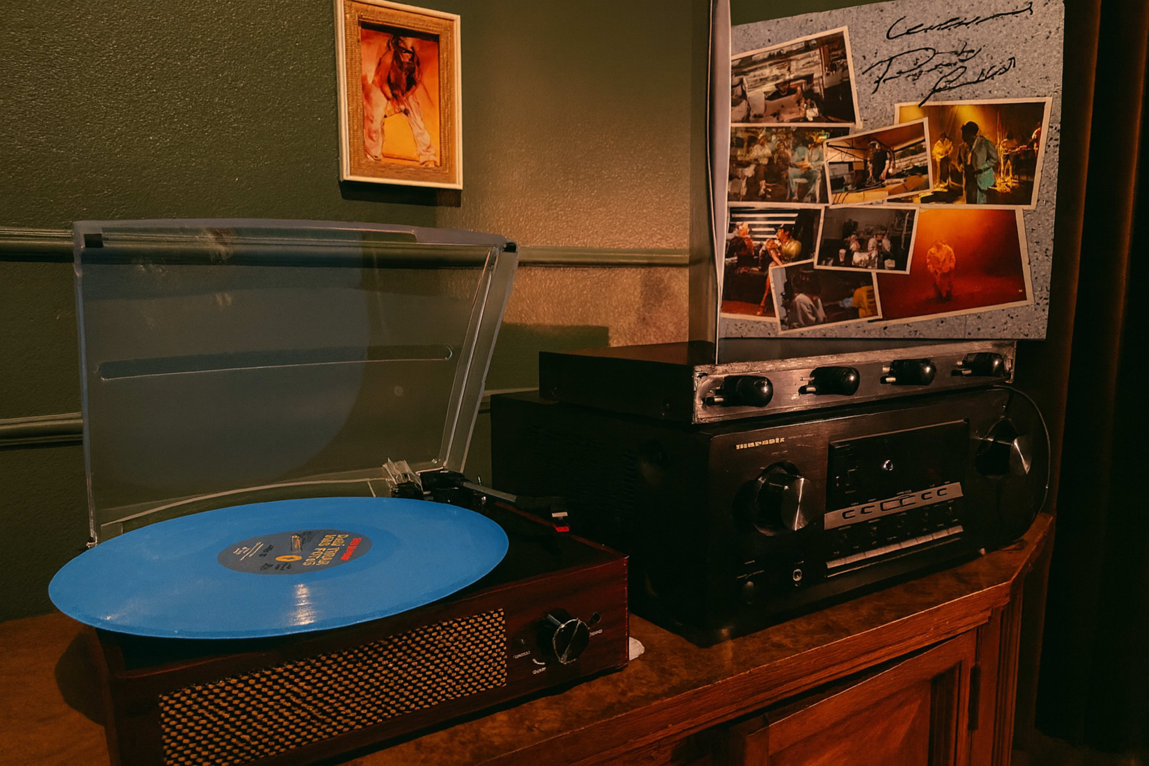The image features a blue vinyl record on a turntable next to a black audio receiver, with a framed photo and a collage of images on the wall behind.